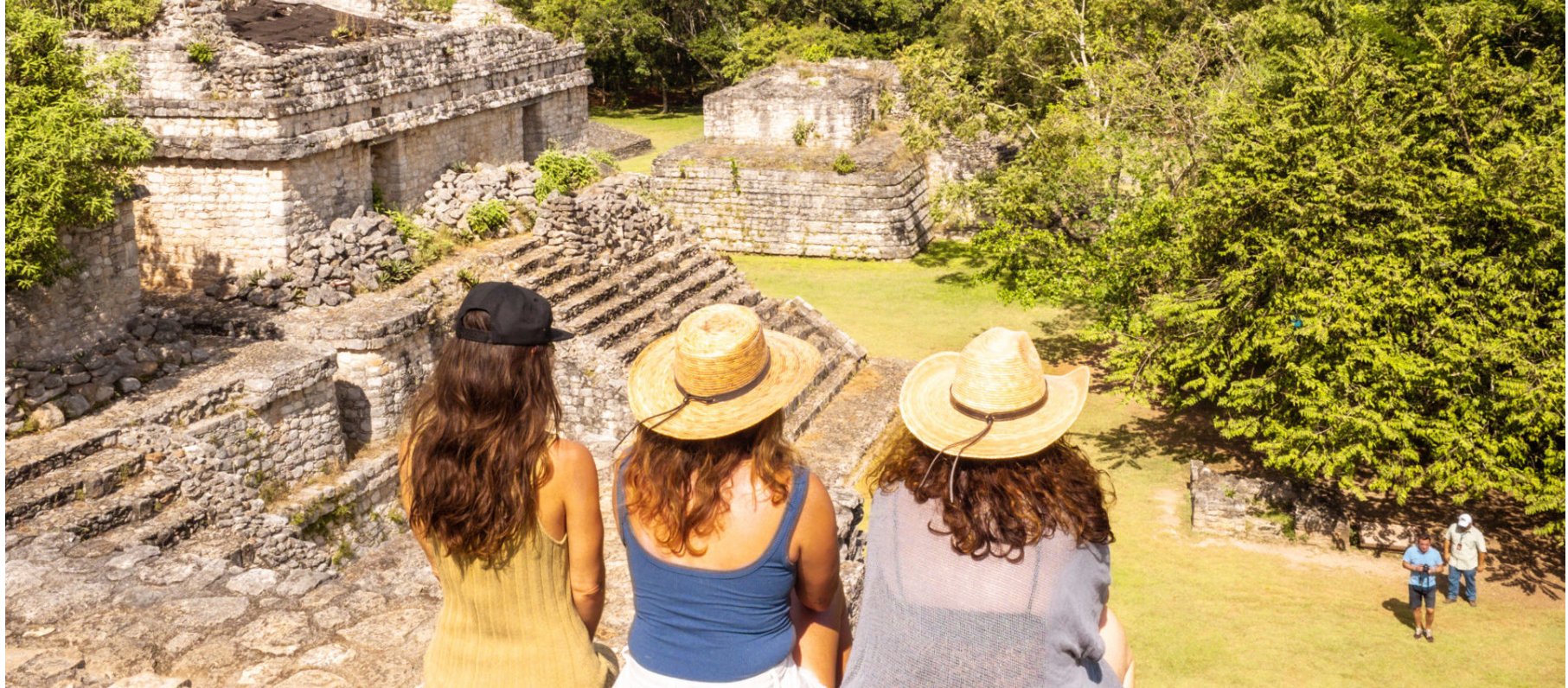 Three women visiting ancient Mayan ruins, wearing hats and casual clothing, seated on a stone ledge, looking at the stepped stone structures and lush green trees.