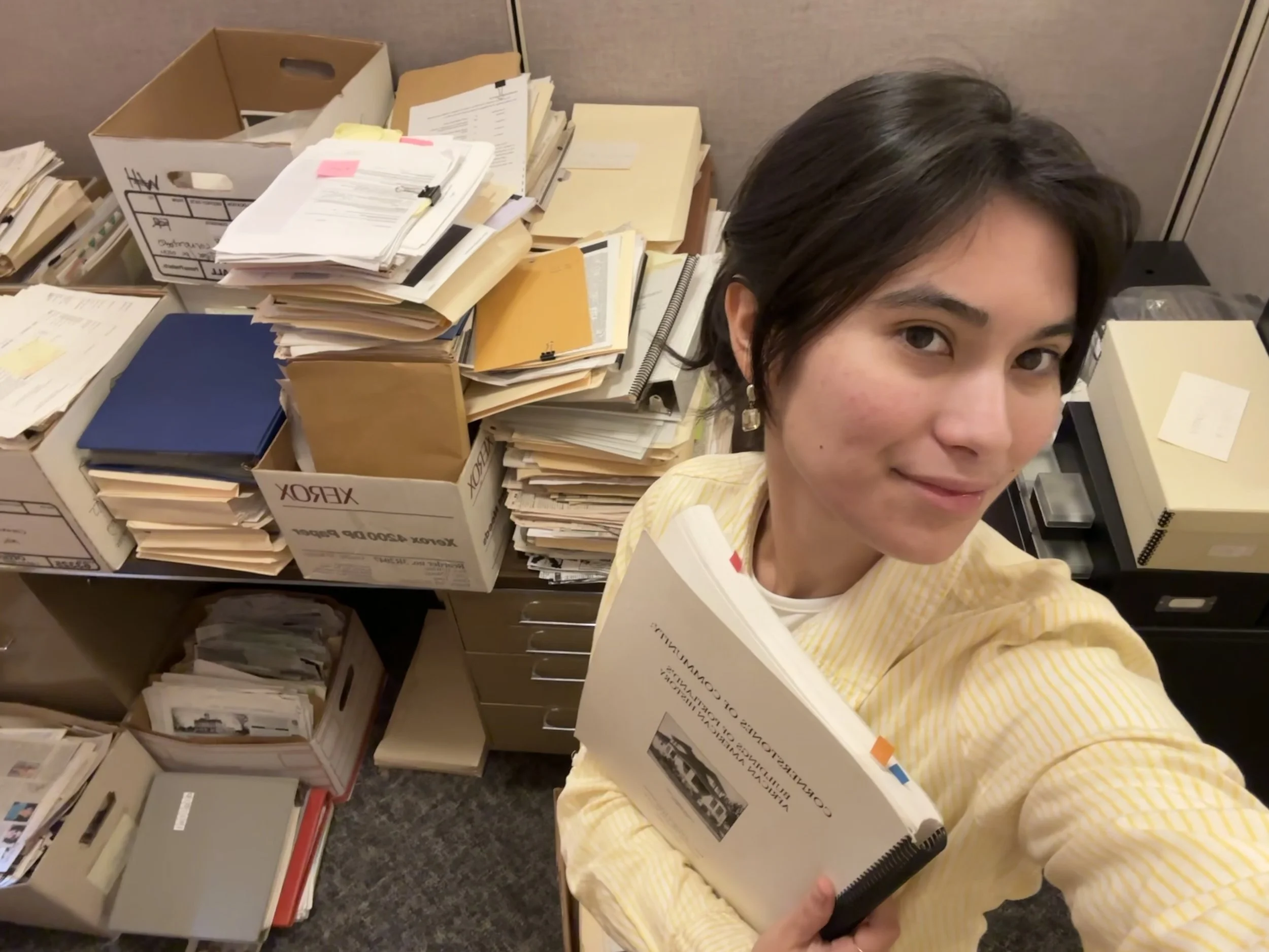 Moya Moses takes a selfie in front of some of the documents used to make Cornerstones of Community