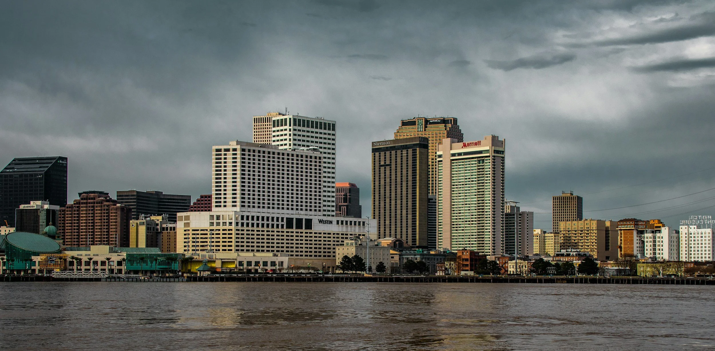 City skyline with various high-rise buildings under dark, cloudy sky, seen from across a body of water.