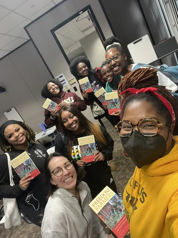 A diverse group of women smiling and holding brochures titled 'Freedom Farmers' in an indoor setting, some wearing glasses and masks.