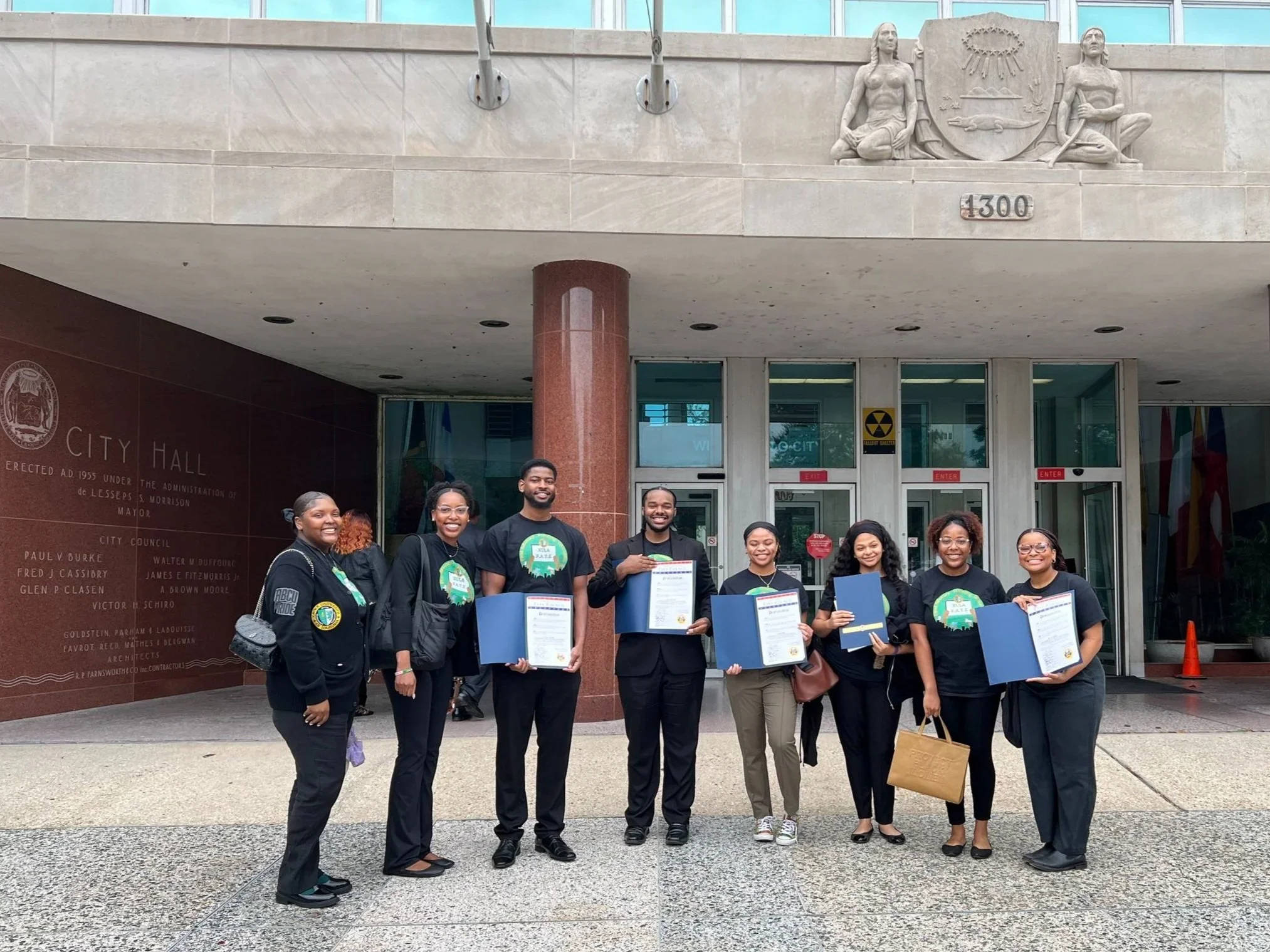 Group of eight people standing in front of a city hall, holding certificates and smiling, with city hall entrance behind them.