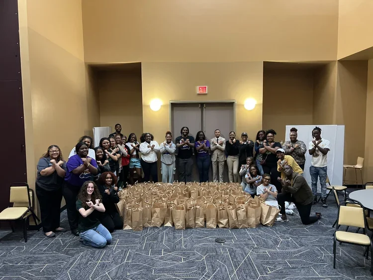 Group of people gathered in a large room, standing behind a table with brown paper gift bags, all making an 'X' with their arms crossed.