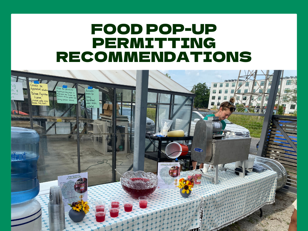 A food pop-up stand serving drinks, with a large water jug, small cups filled with red beverage, and decorative sunflower vases on a table. A woman is operating a machines behind the table, with signs and notes attached to the structure nearby.