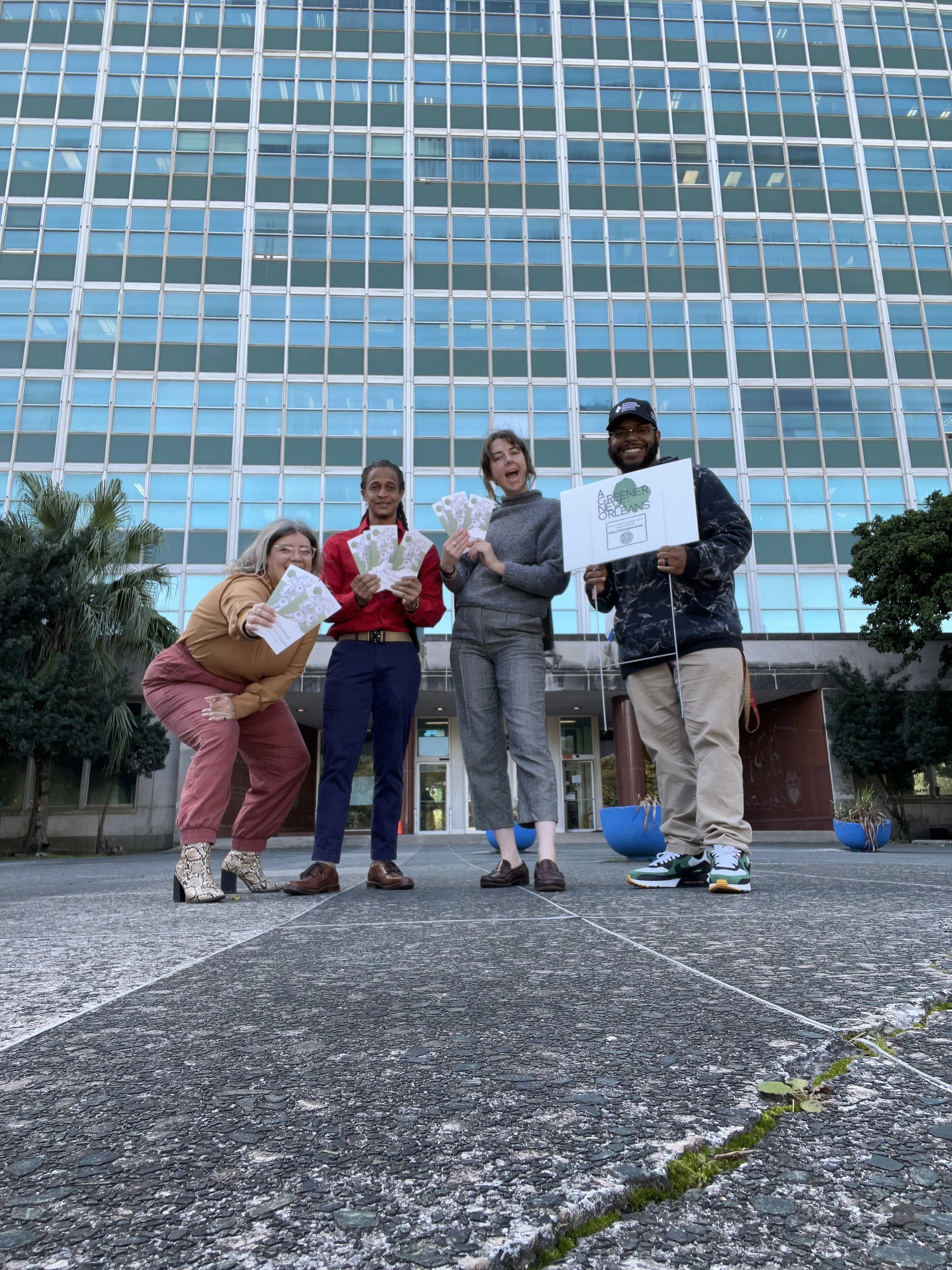 Four diverse people standing outside a modern glass building, smiling and holding posters or flyers, with a cityscape background.