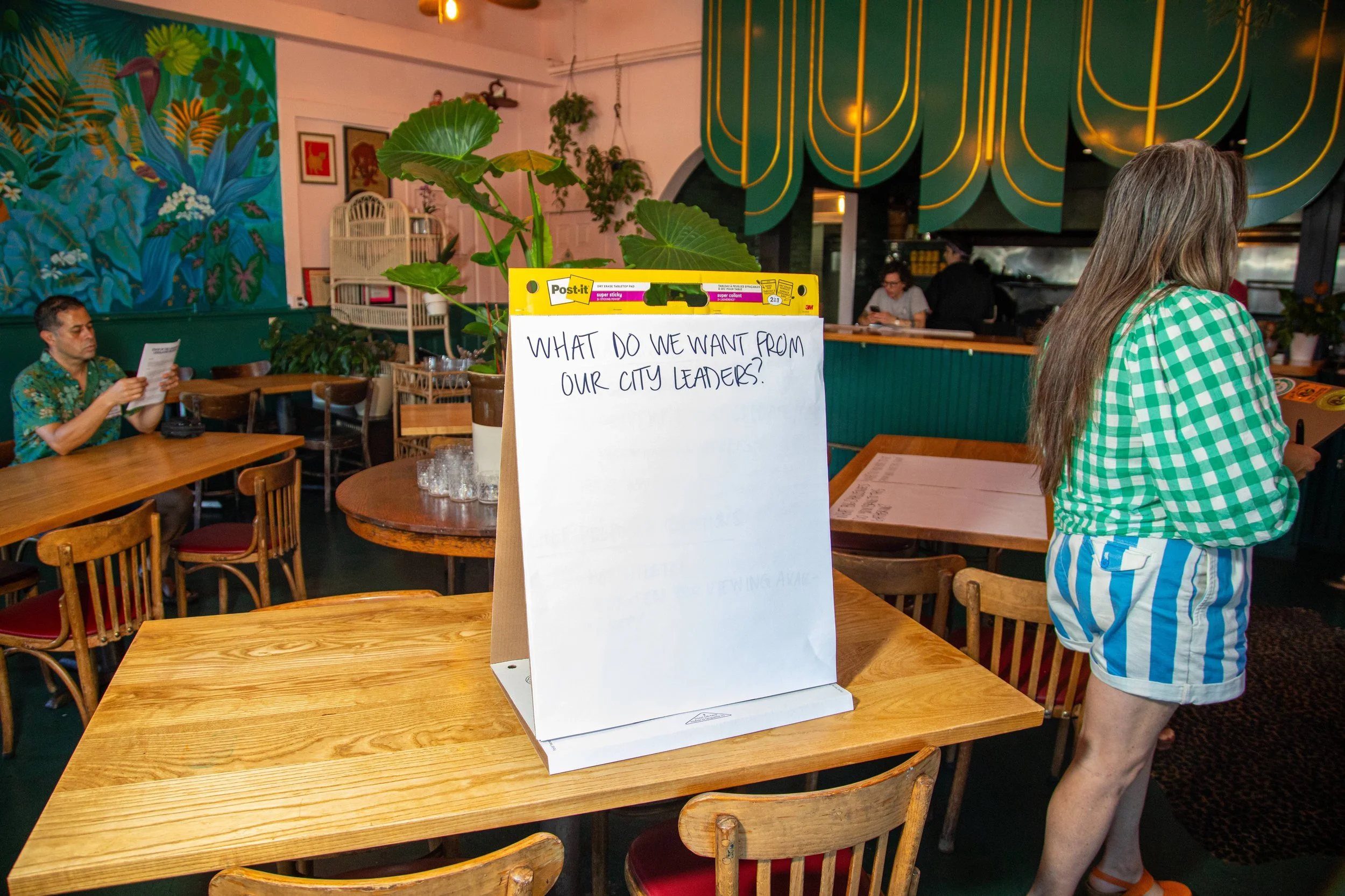 Interior of a restaurant with a large table in the foreground holding a sign that reads 'What do we want from our city leaders?' A woman with long hair, wearing a green and white checkered shirt and striped shorts, stands near the table. Other diners are seated at tables in the background, and the decor includes jungle-themed mural art and green lighting.
