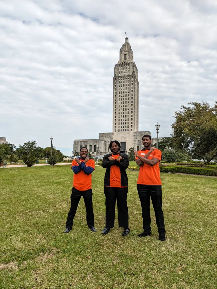 Three men standing on a grassy area in front of the Empire State Building, all crossing their arms and smiling at the camera. They are wearing matching orange and navy blue sports shirts.