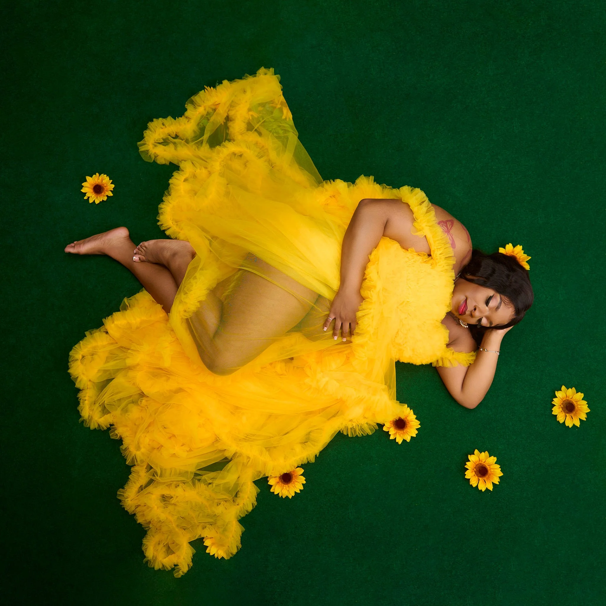 Pregnant woman in a yellow tulle dress lying on a green background with sunflowers