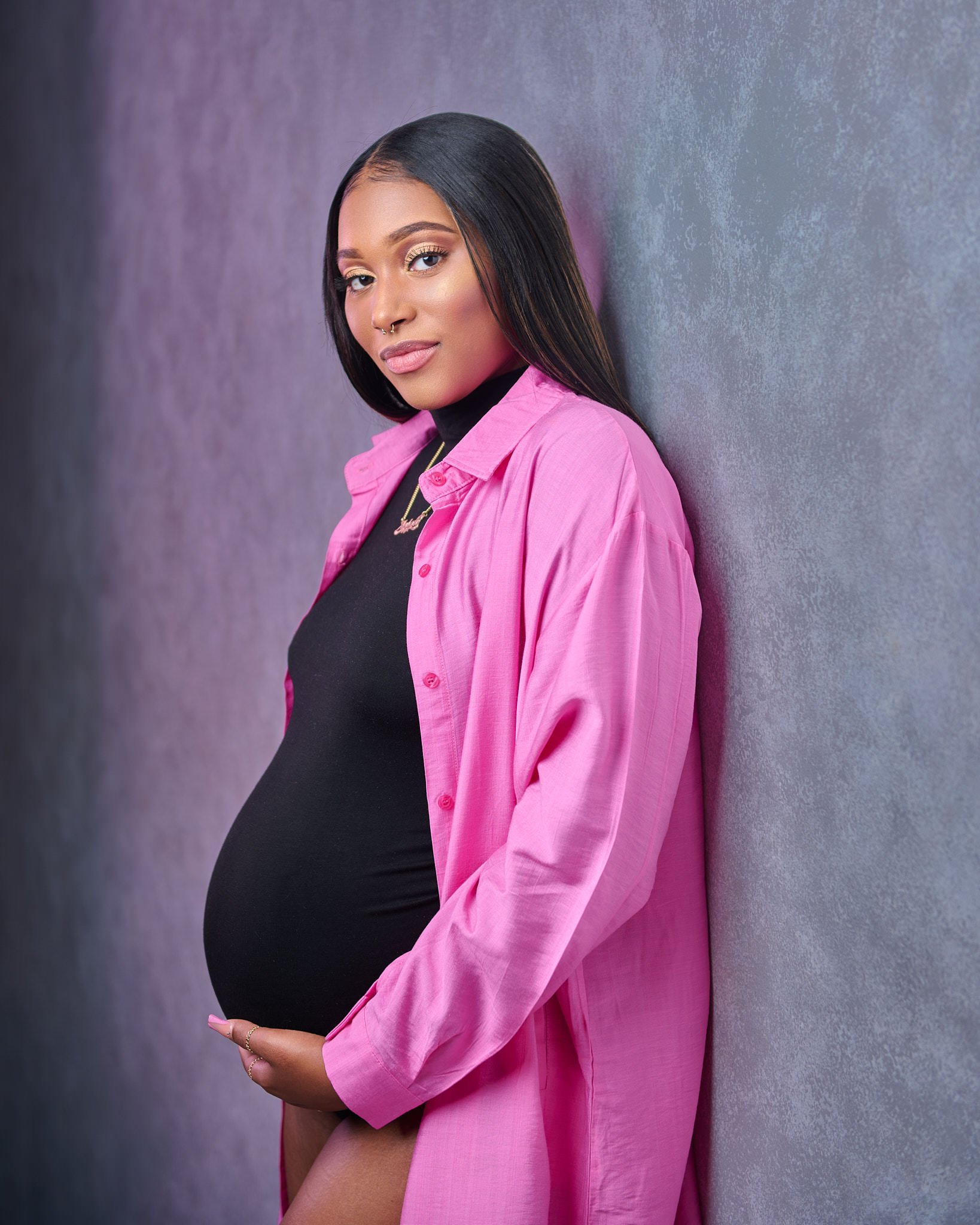 Pregnant woman in a black dress and pink shirt against a gray background