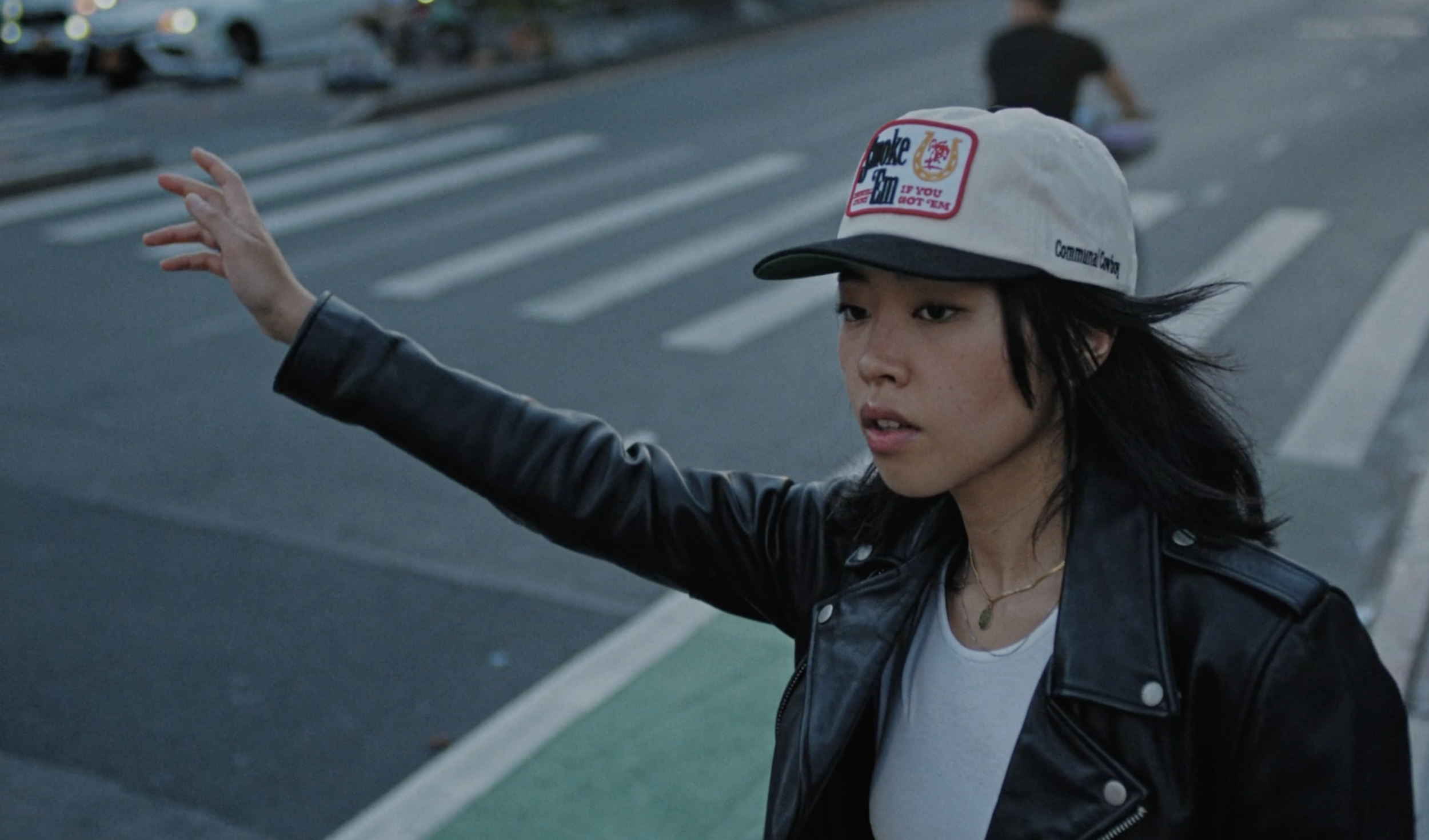 A woman with black hair wearing a baseball cap, a black leather jacket, and a white t-shirt stands on a crosswalk, extending her arm as if to signal or stop traffic.