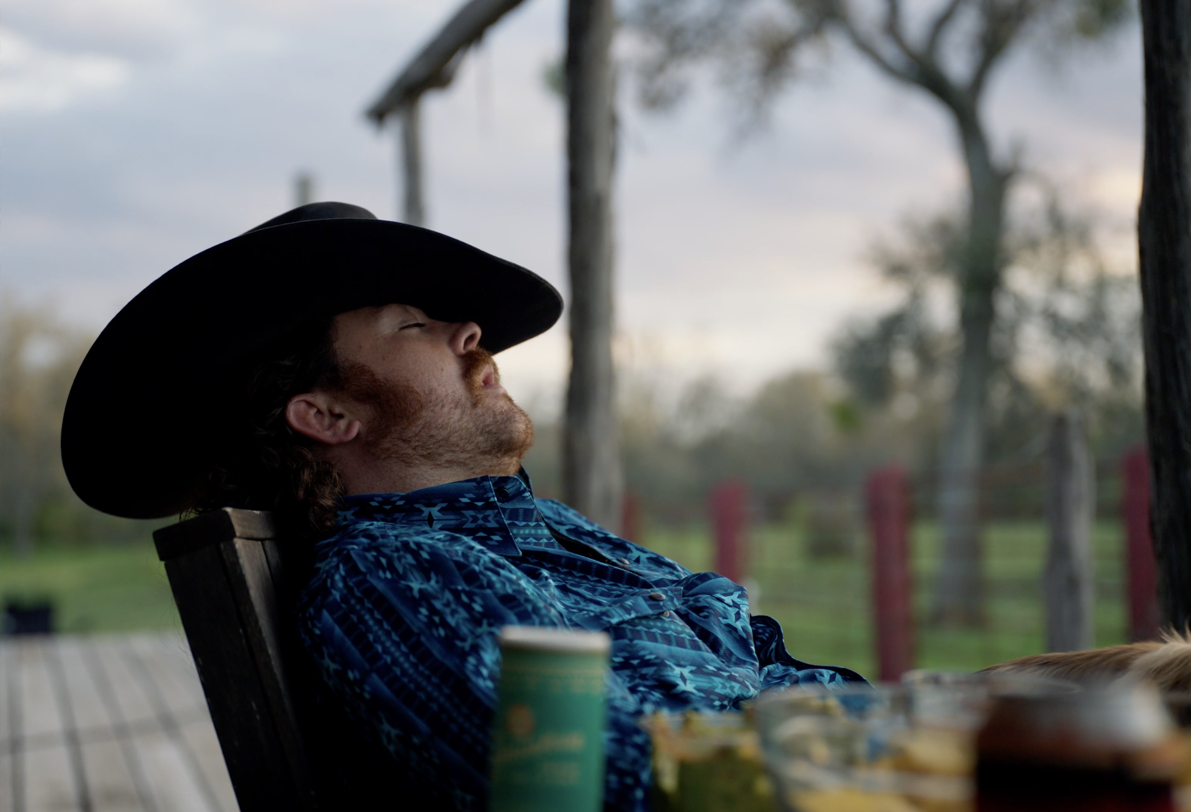 A man with a beard and long hair, wearing a blue patterned shirt and a black cowboy hat, is sitting outdoors with his eyes closed, leaning back in a chair as the sun sets behind trees.