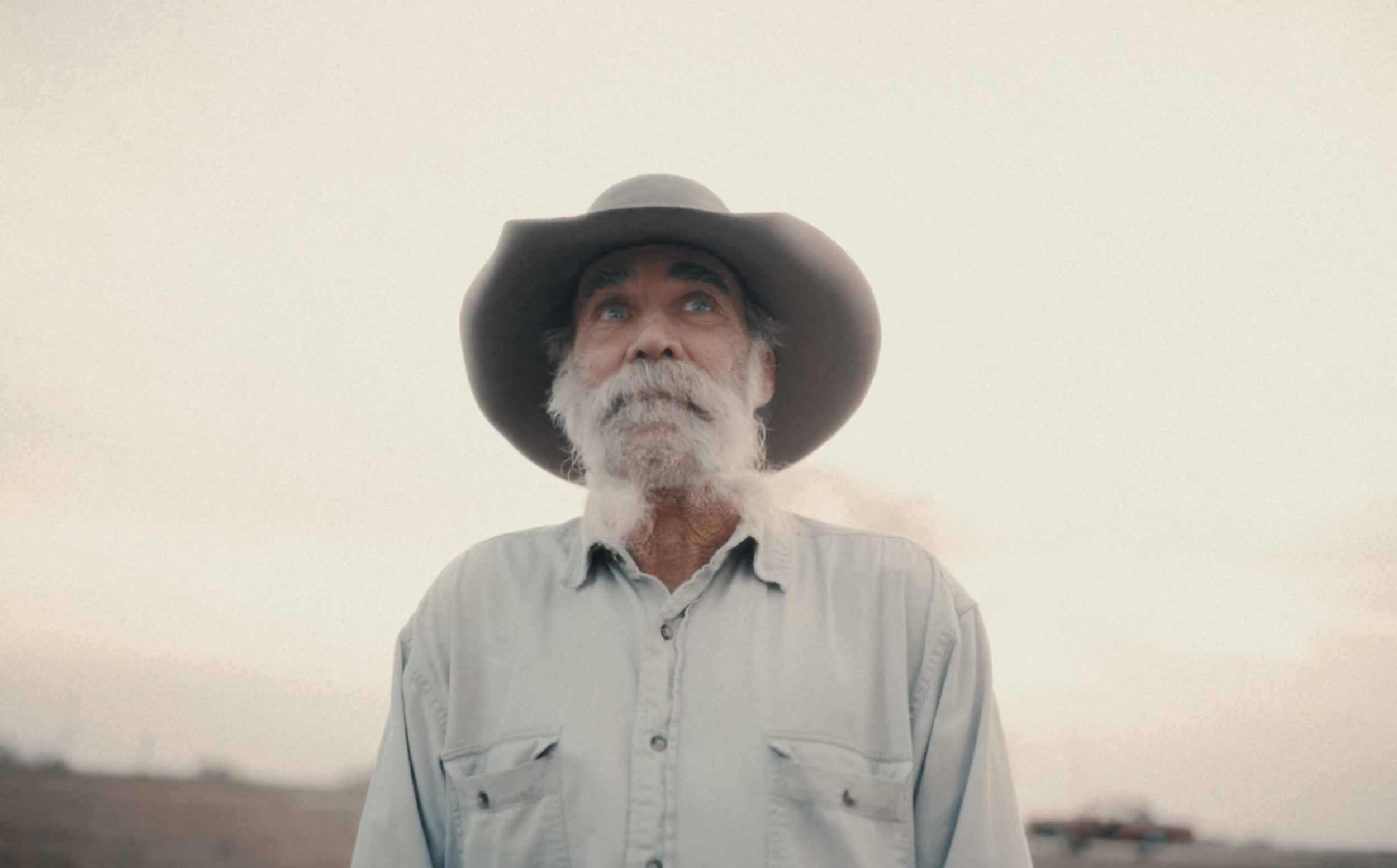 Older man with gray beard and blue eyes, wearing a wide-brimmed hat and light-colored shirt, looking upward against a bright sky.