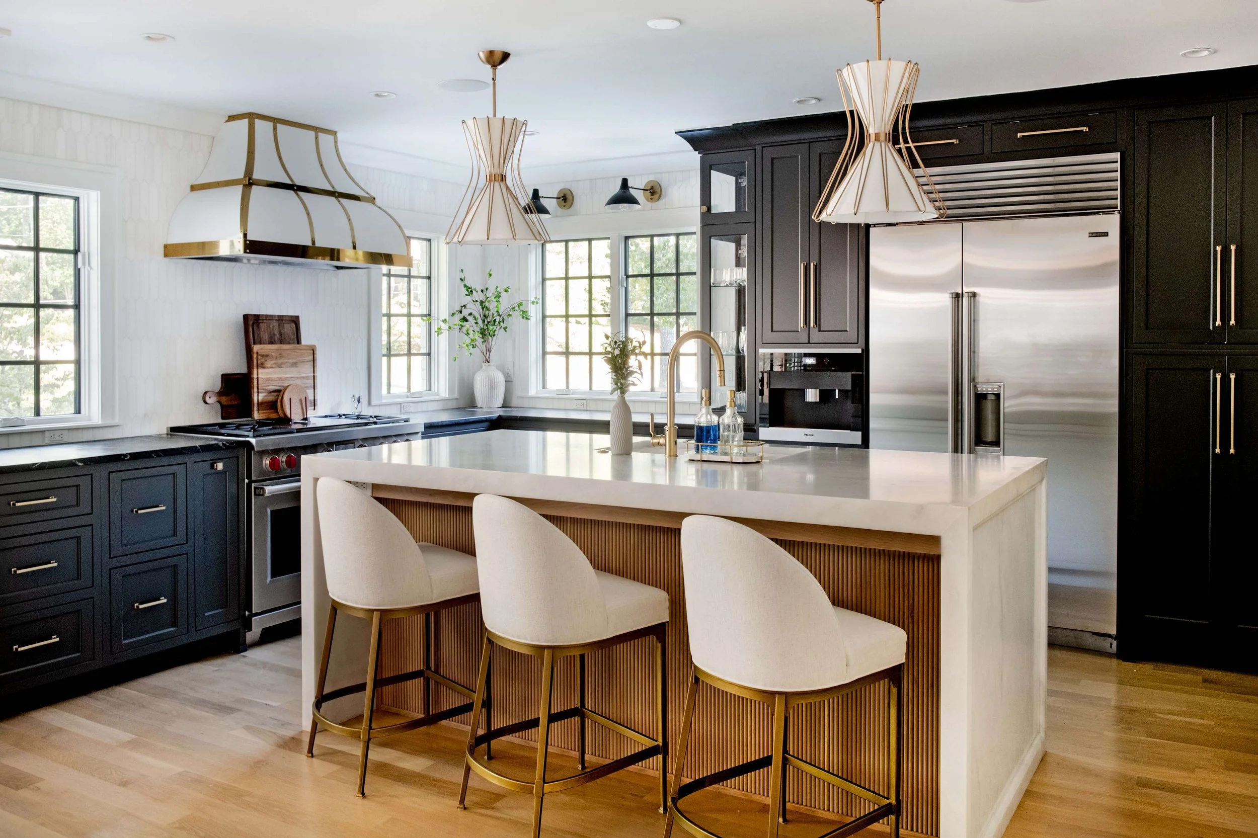 A modern kitchen with black cabinets, stainless steel refrigerator, white island with bar seating, pendant lights, and natural light from large windows.