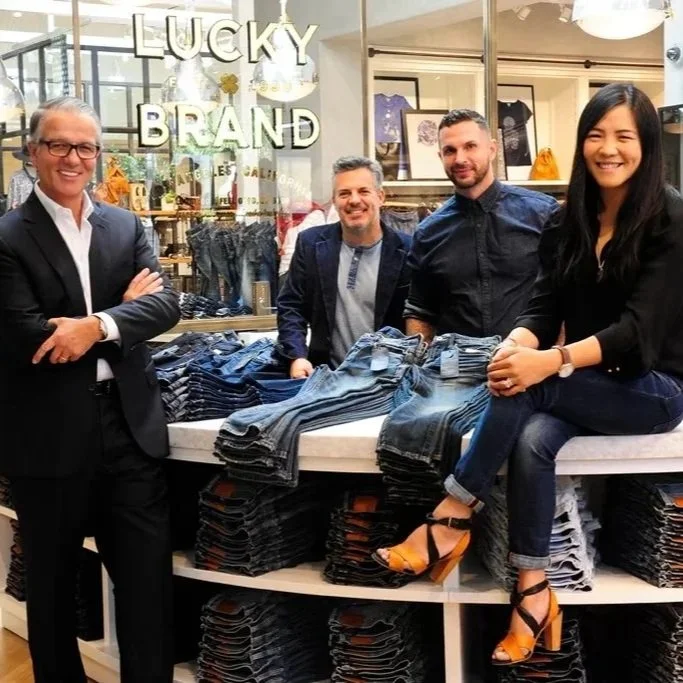 Four people standing behind a display of jeans at a Lucky Brand store, smiling at the camera.