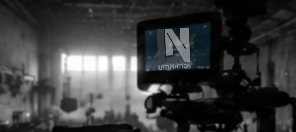 A camera on a tripod in a dim, industrial-style space with large windows, showing a screen with a stylized 'N' and the text 'LATIN NATION'.