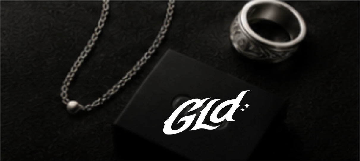 A silver chain necklace with a pearl pendant, a black box with the word 'Gld' in white letters, and a decorative ring with black and silver patterns, on a black textured surface.