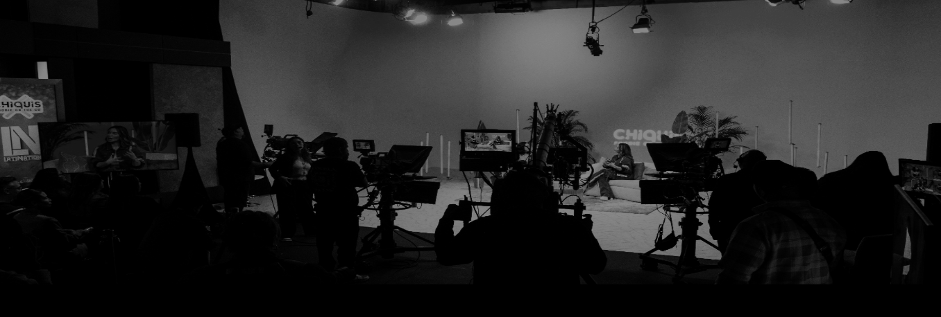 A behind-the-scenes view of a television studio with cameras, crew members, and a host or guest seated on a couch, set with plants and backdrop.