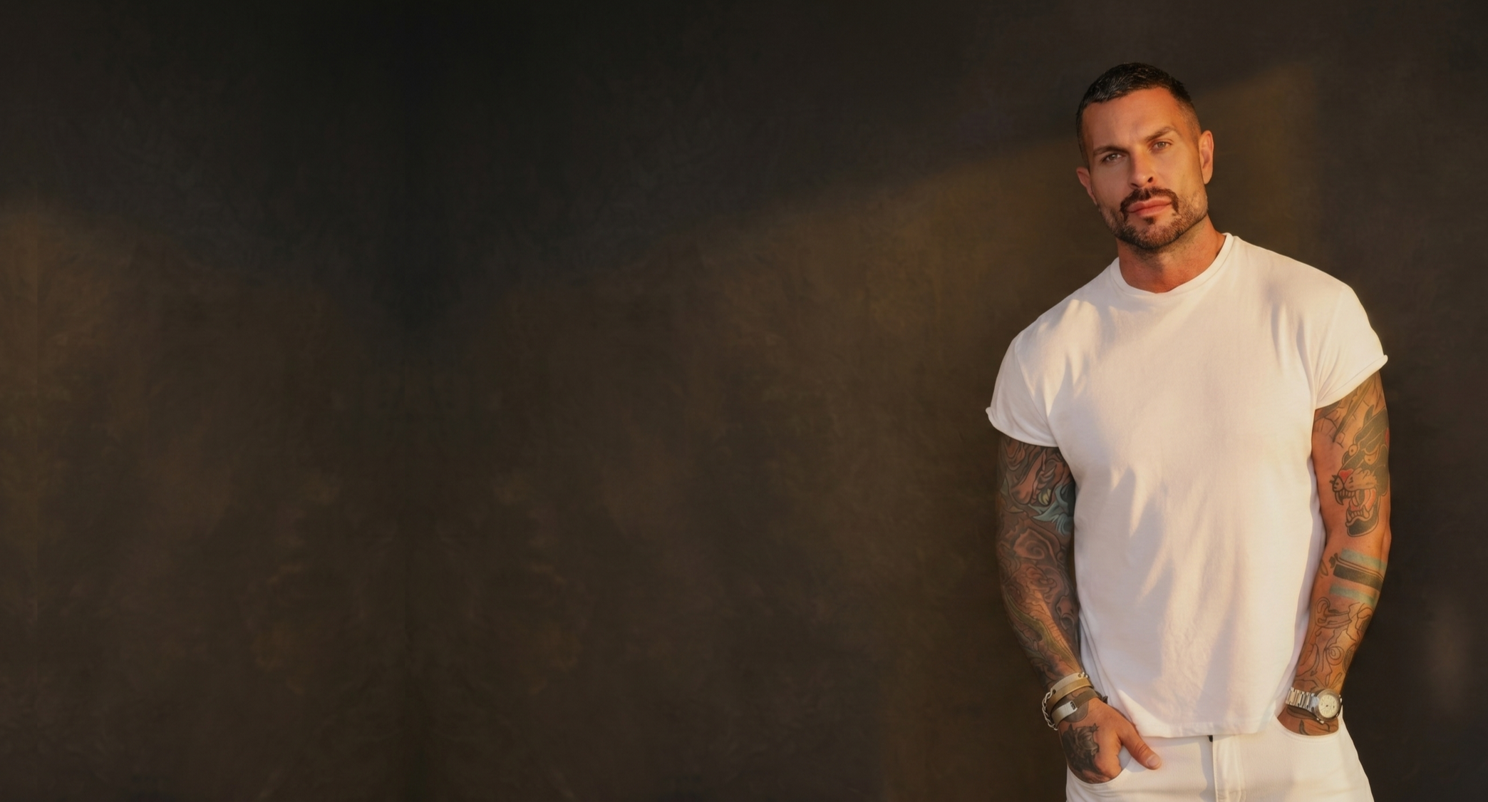 A man with short dark hair and a beard, wearing a white T-shirt, has colorful tattoos on his arms, and is standing against a dark background.