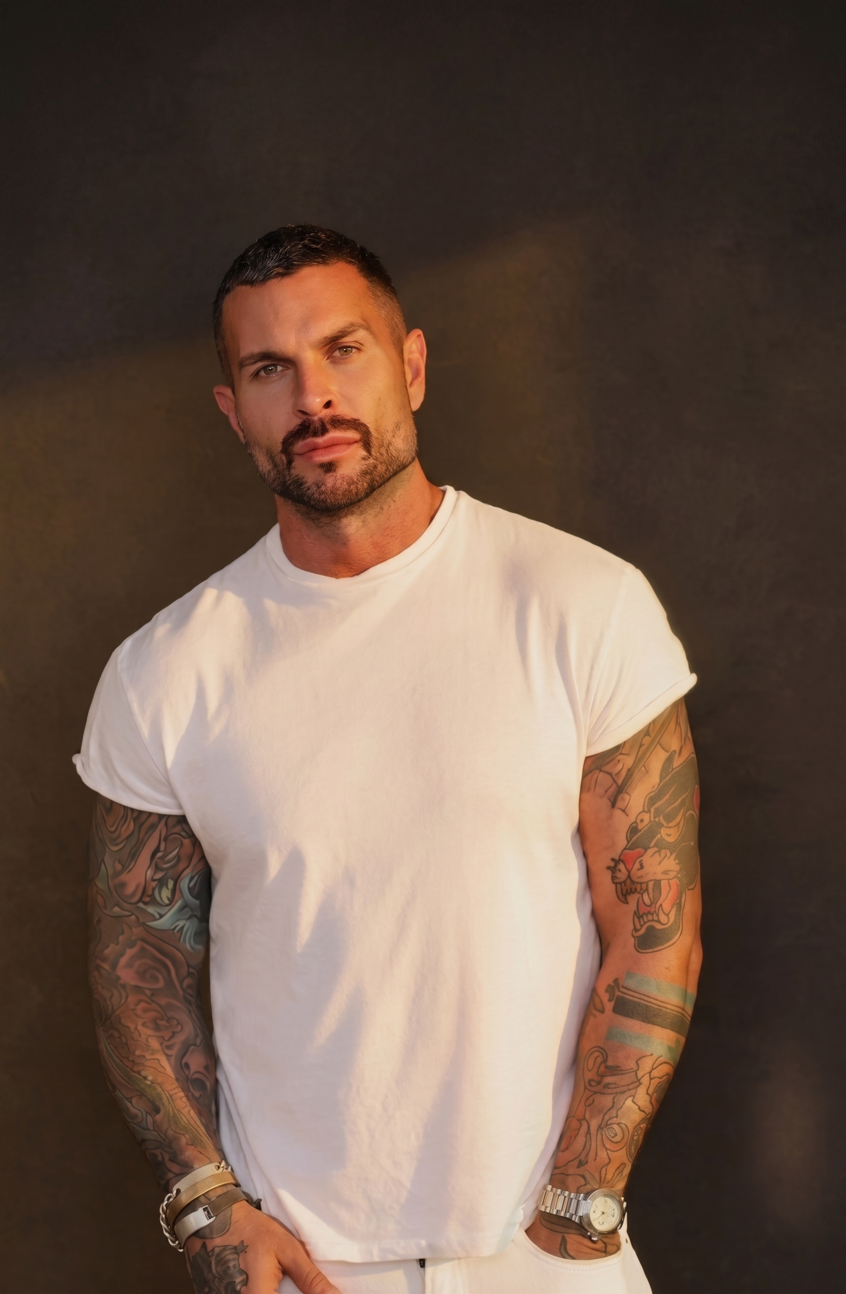 A man with tattoos on both arms, short dark hair, and a beard, wearing a white t-shirt, standing against a dark background.