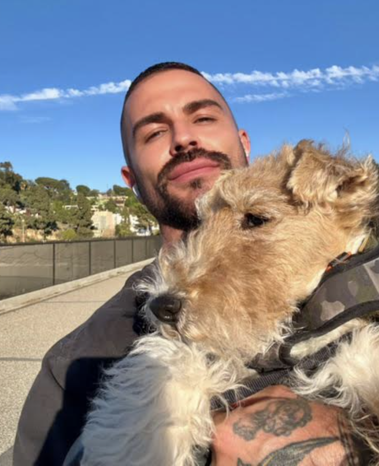 A man with a beard and tattoos holding a tan and white dog outdoors on a sunny day with a clear blue sky.