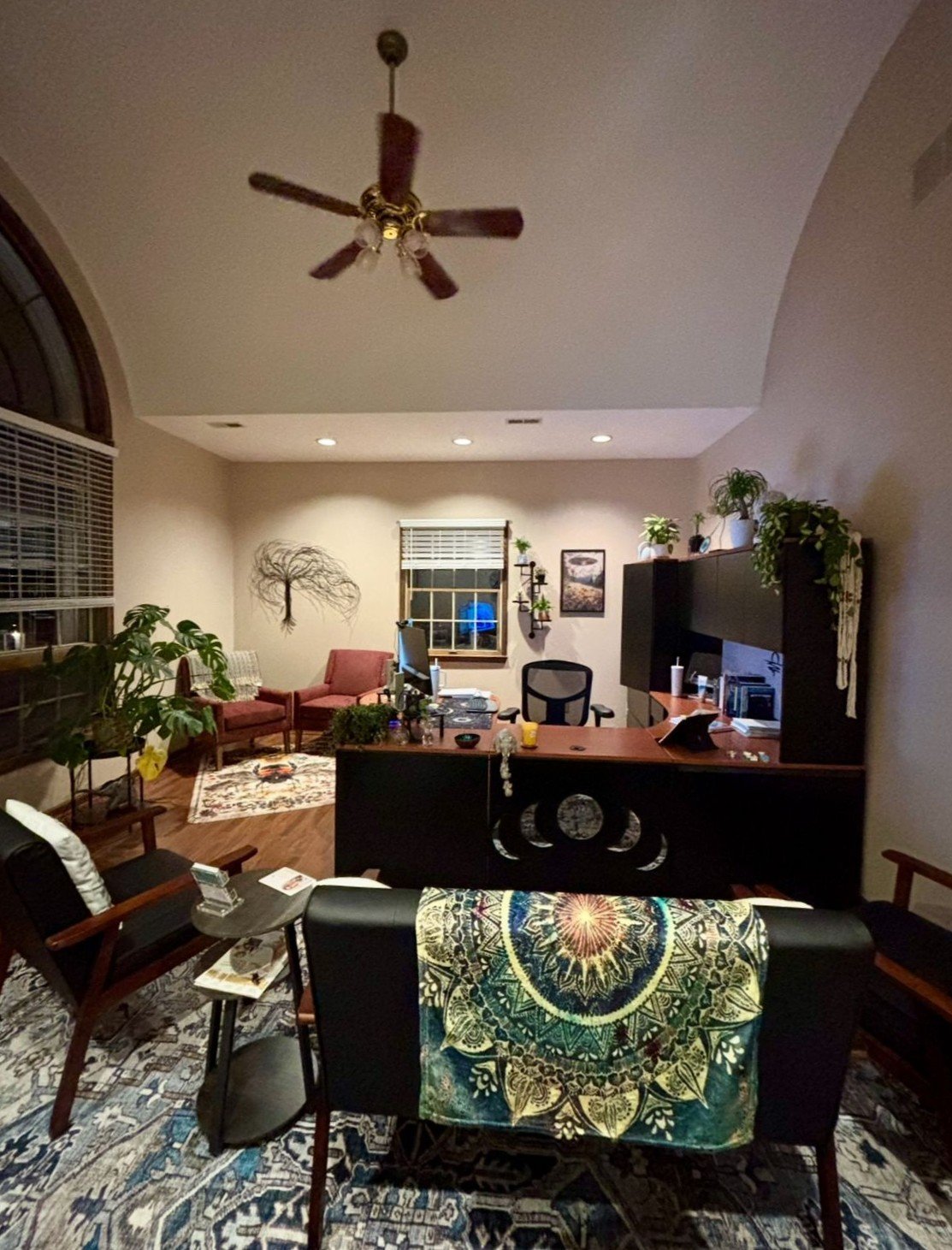 Interior view of a cozy office with a large wooden desk, black office chair, and various plants. There are couches and chairs in the background, a window, wall art, and a ceiling fan.