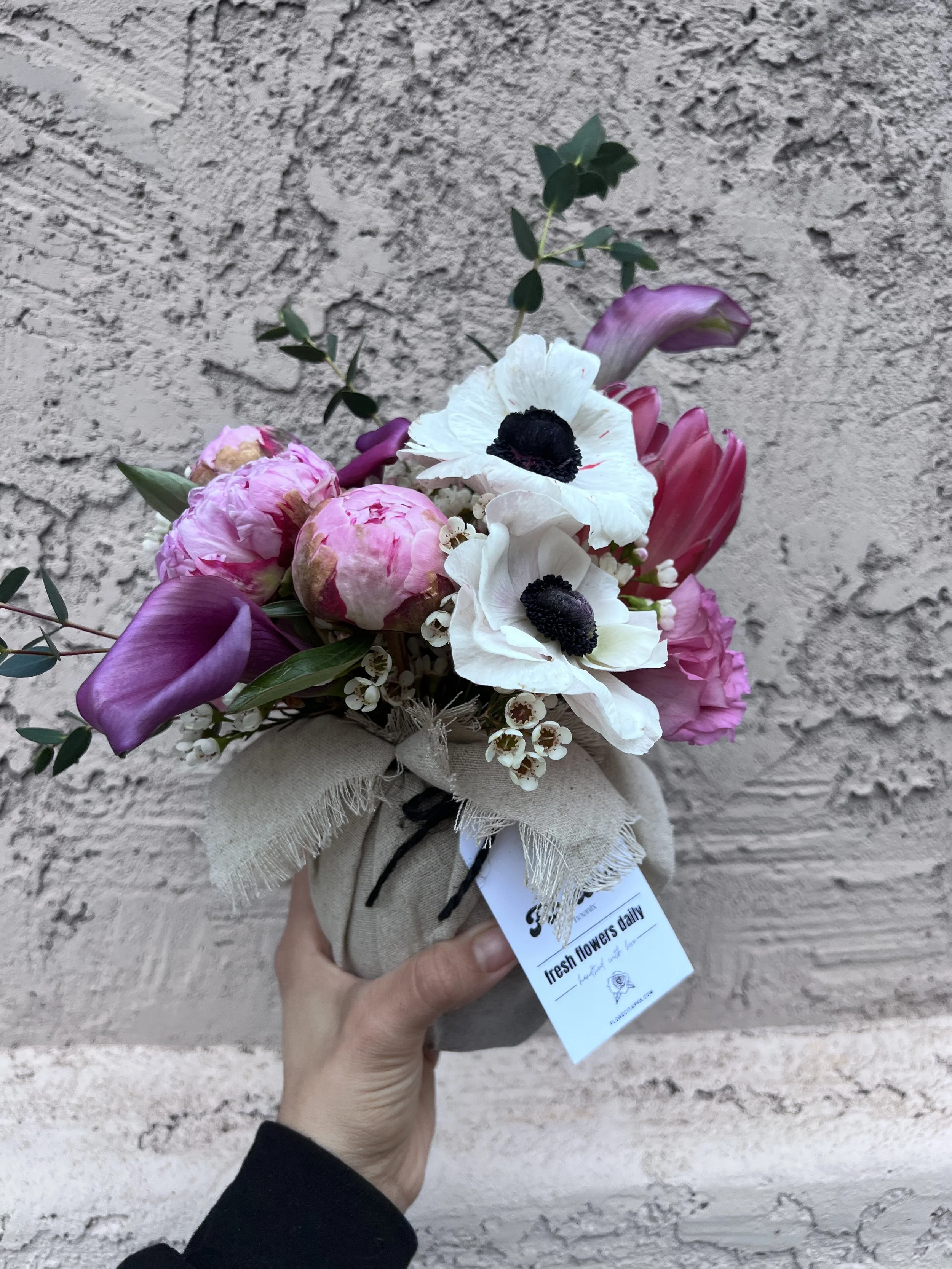 A bouquet of pink peonies, white poppies, purple tulips, and greenery wrapped in burlap with a label that reads "fresh flowers daily."