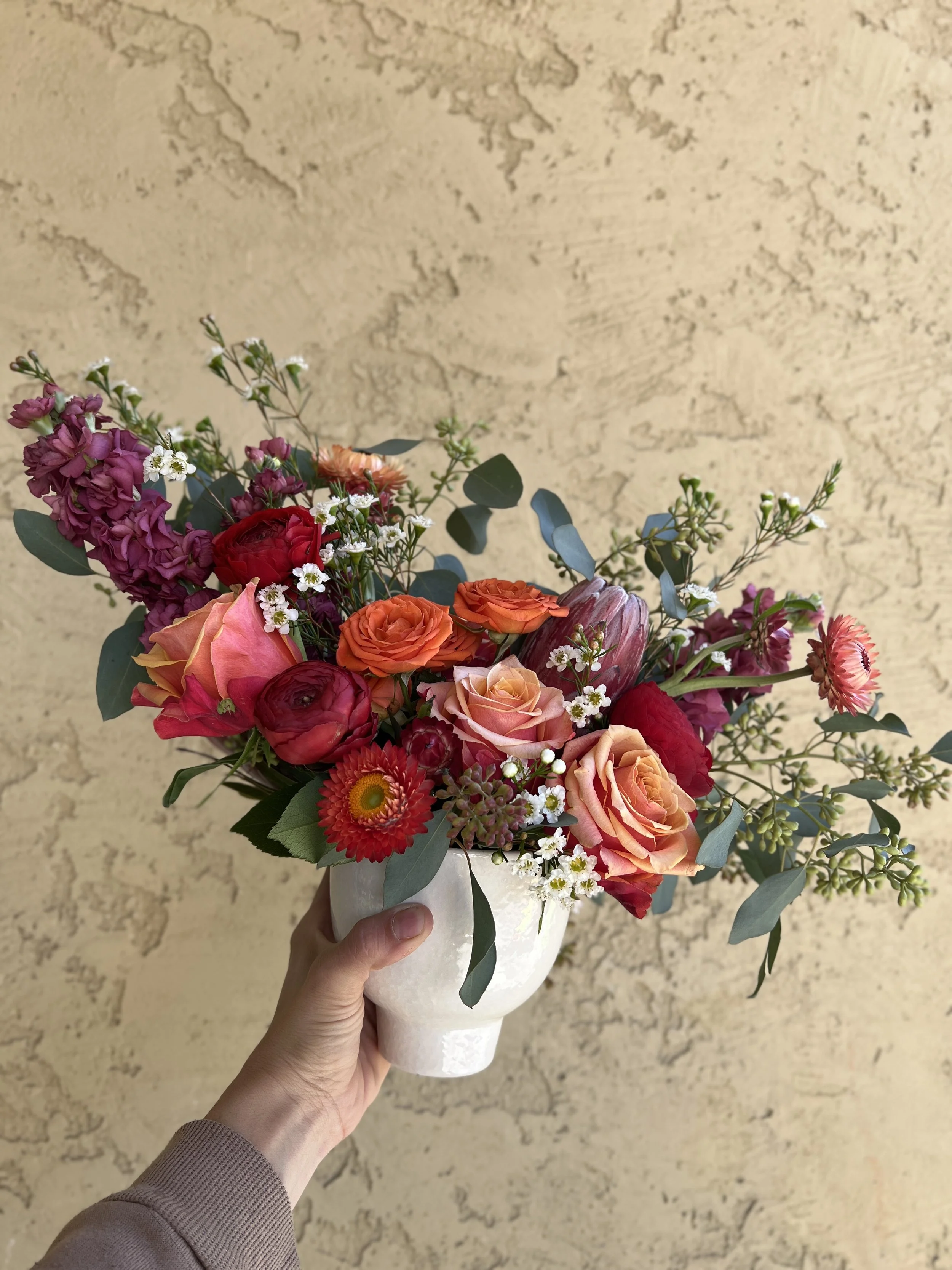 A hand holding a white vase with a colorful bouquet of flowers, including roses, daisies, and other mixed blooms against a textured tan wall.
