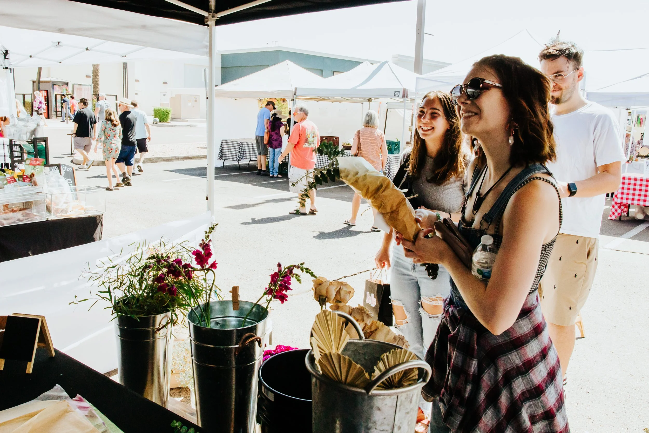 Three young women are shopping at an outdoor market stall, with one holding a bouquet of flowers and another holding a bottle of water. They are smiling and appear to be enjoying their visit. Behind them, other market stalls and people are visible.