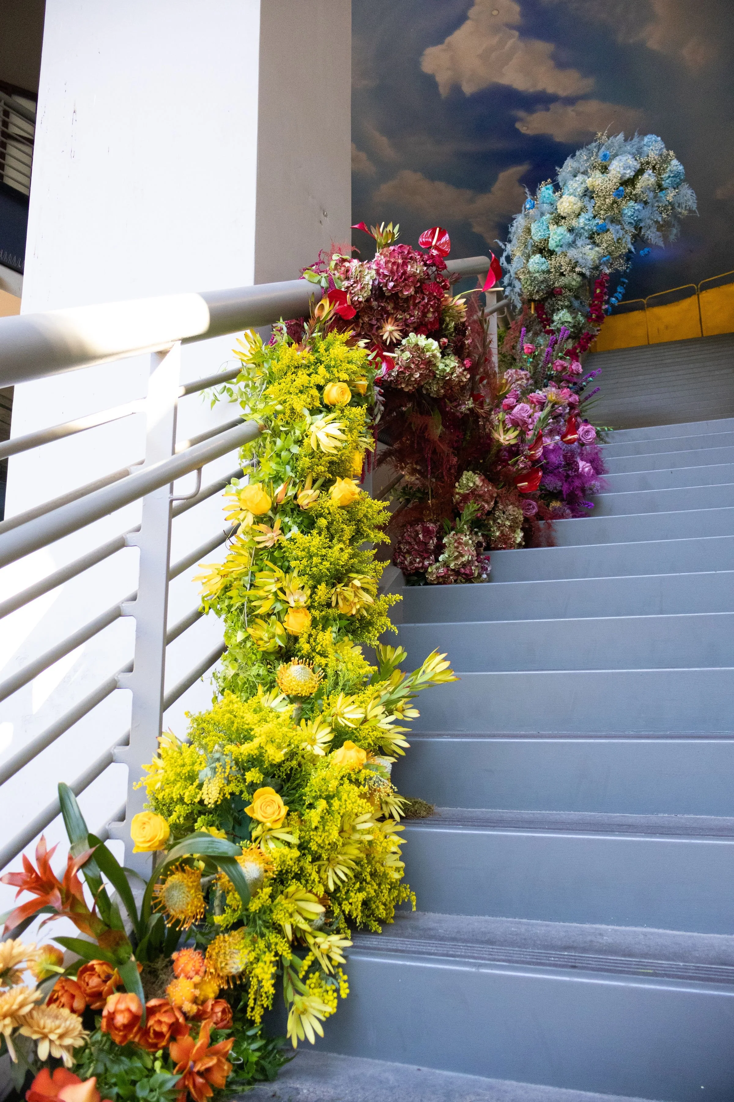 Colorful flower arrangements placed along a staircase railing, with a painted sky mural on the ceiling in the background.