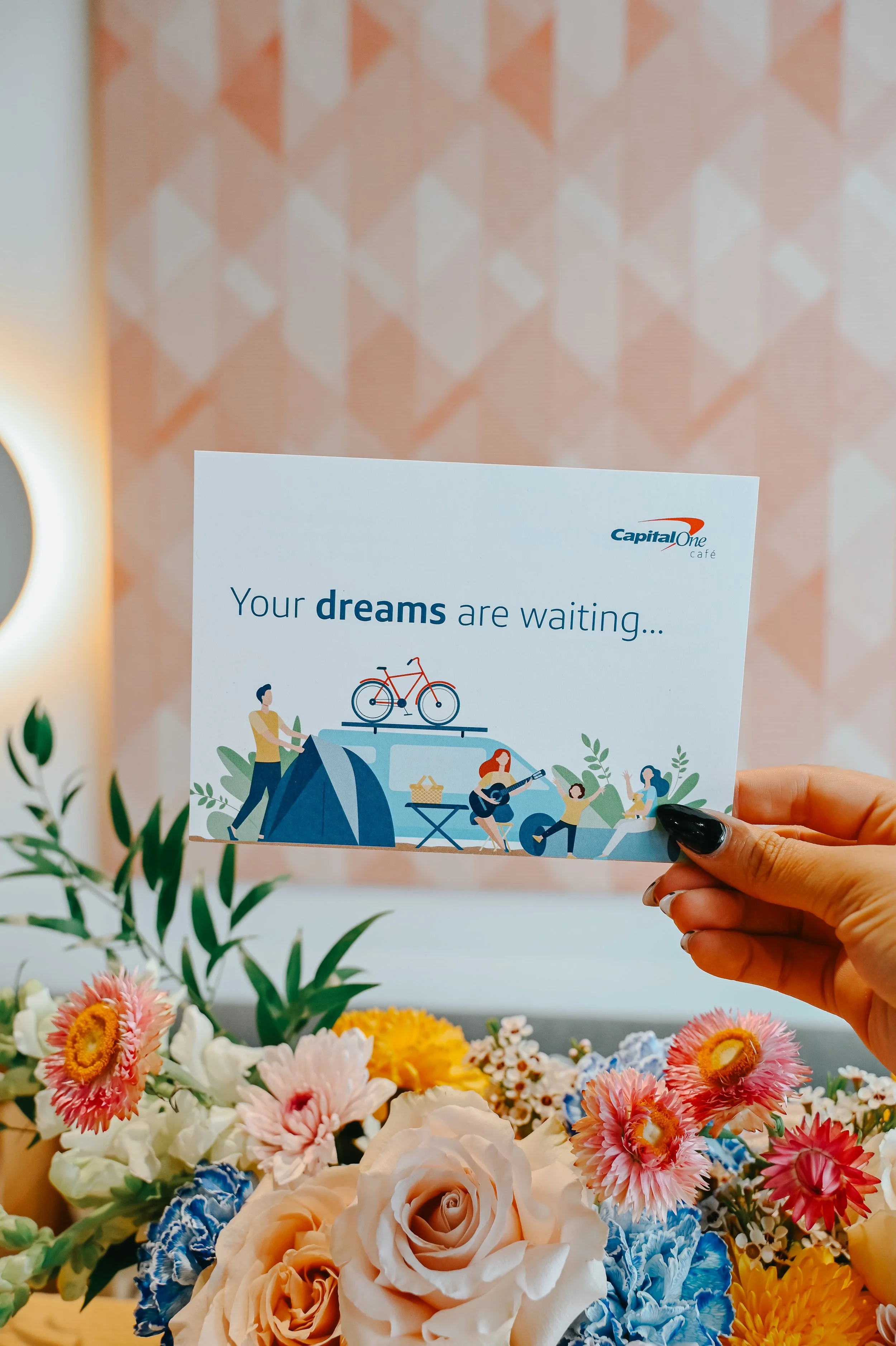 A hand holding a promotional card with the message 'Your dreams are waiting...' from Capital One Café, with illustrations of people camping, playing guitar, and riding bikes, behind a floral arrangement with pink, white, yellow, and blue flowers.
