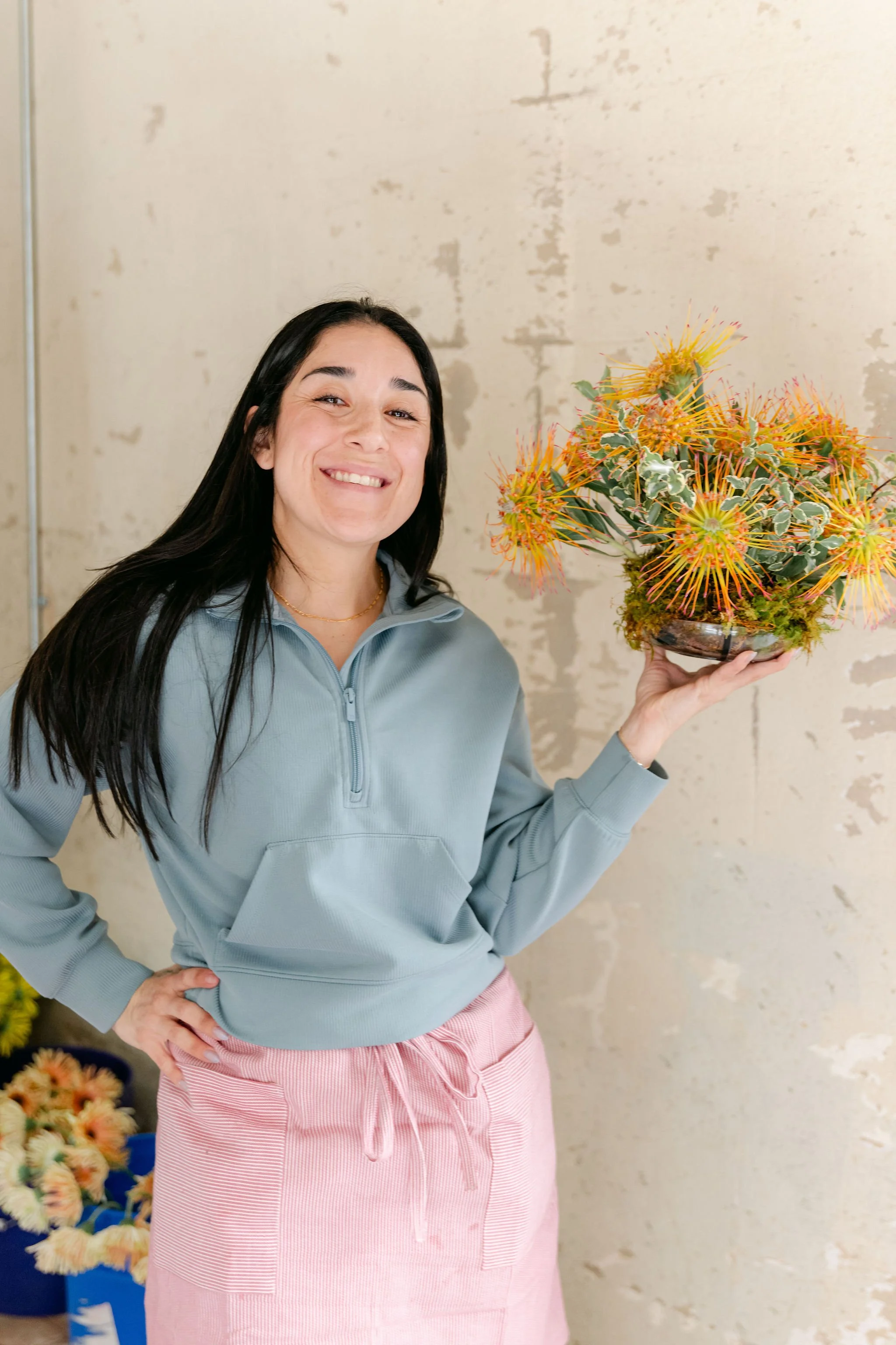 A woman with long dark hair smiling and holding a colorful bouquet of flowers in her right hand, standing against a beige textured wall.