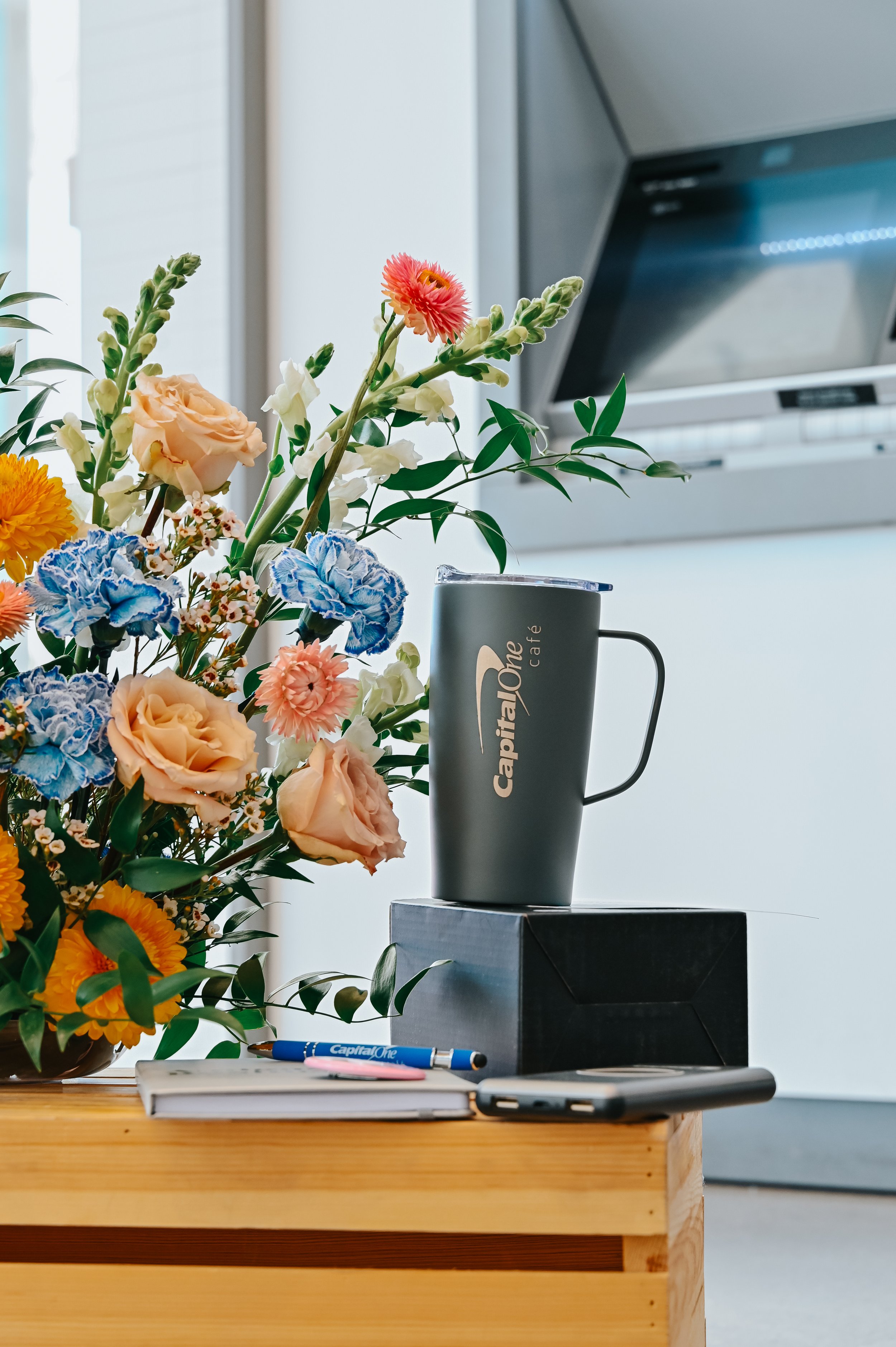 A vase of colorful flowers next to a gray mug with the Capital One logo on a wooden table, with a notepad, pens, and a smartphone in the foreground, and a kitchen range in the background.