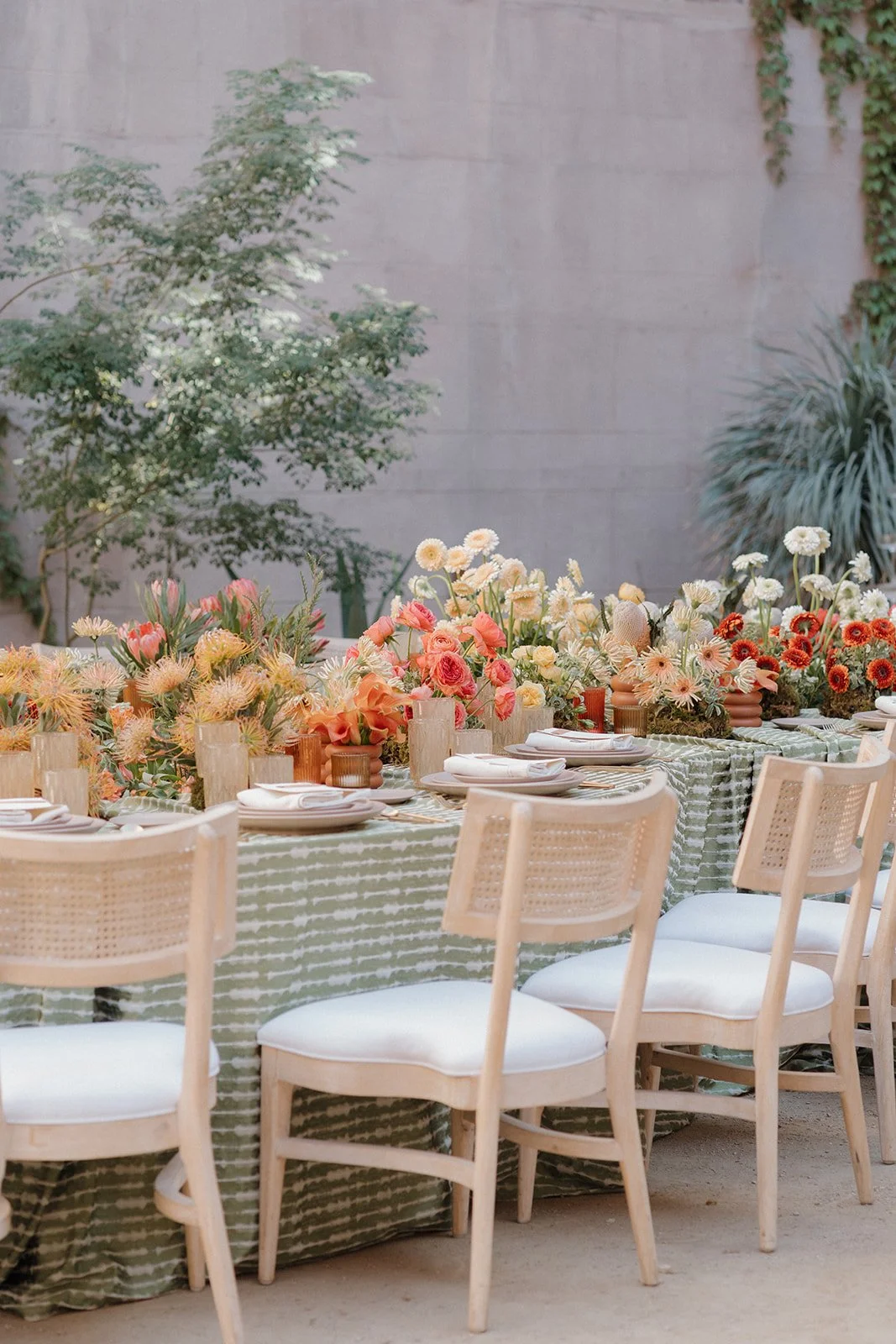 A long rectangular dining table decorated with a green and white striped tablecloth, topped with an elaborate floral centerpiece featuring pink, white, and orange flowers. The table is set with white plates, napkins, and candles. Surrounding the table are light wood chairs with white cushions. In the background, there are large green plants and a concrete wall.