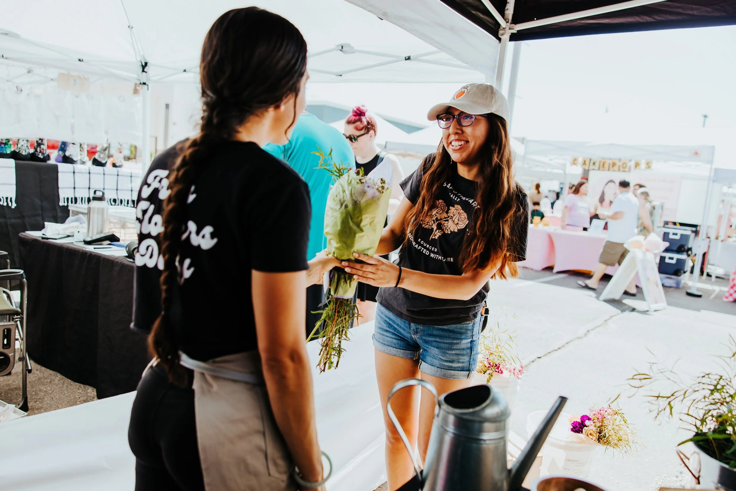 A woman with long hair, glasses, and a gray baseball cap smiling and handing a bouquet of flowers to another woman at an outdoor market or festival.