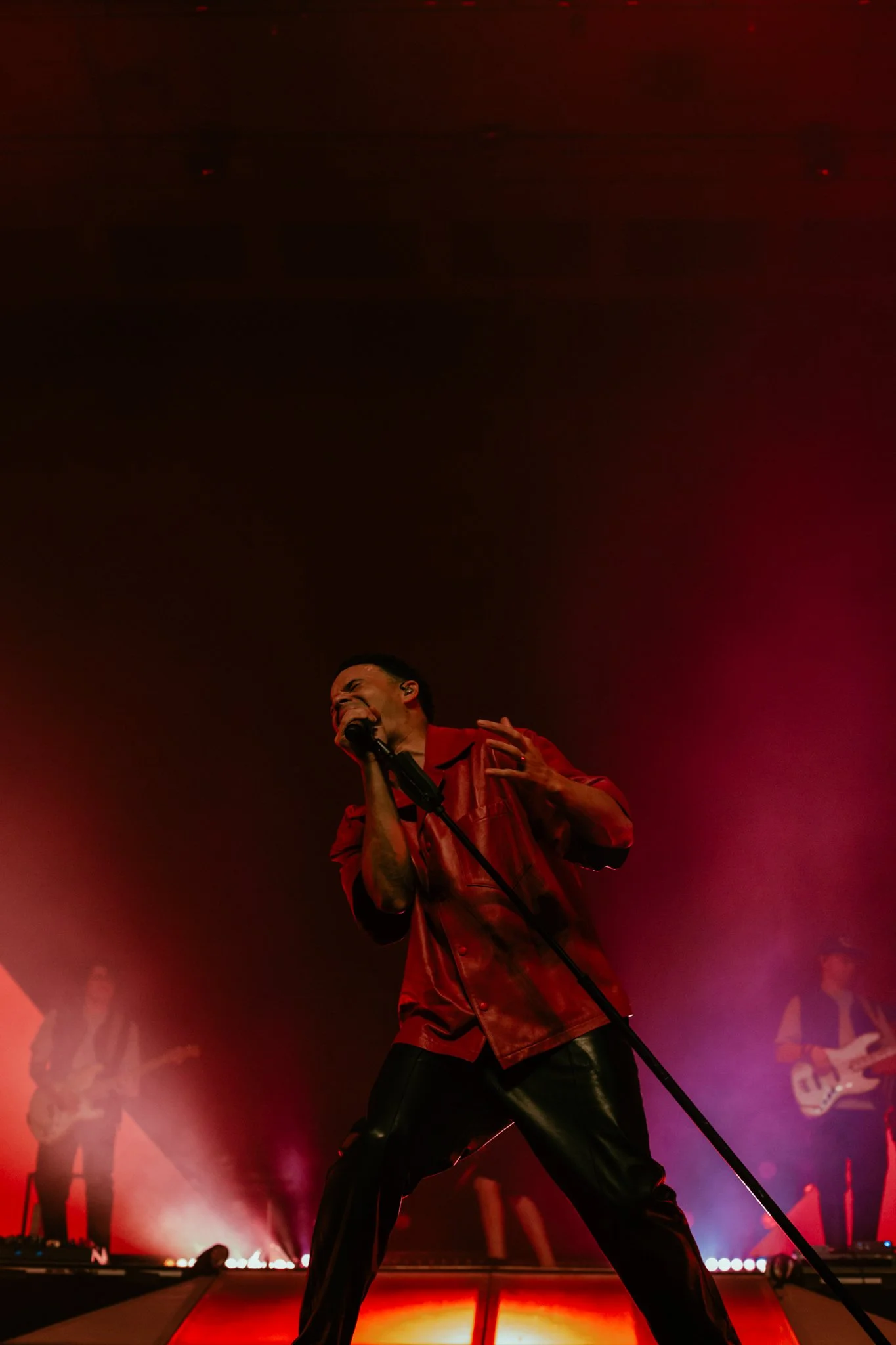 Performer singing passionately on stage, wearing a red shirt and black pants, with a microphone, with band members playing guitars in the background, colorful stage lighting.