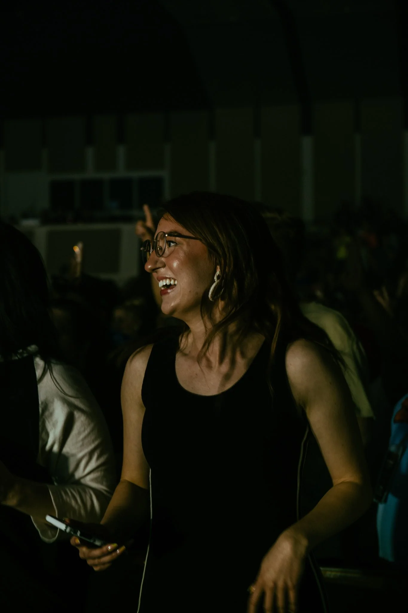 A woman with glasses and earrings smiling at an indoor event, holding a smartphone, in a dark environment.