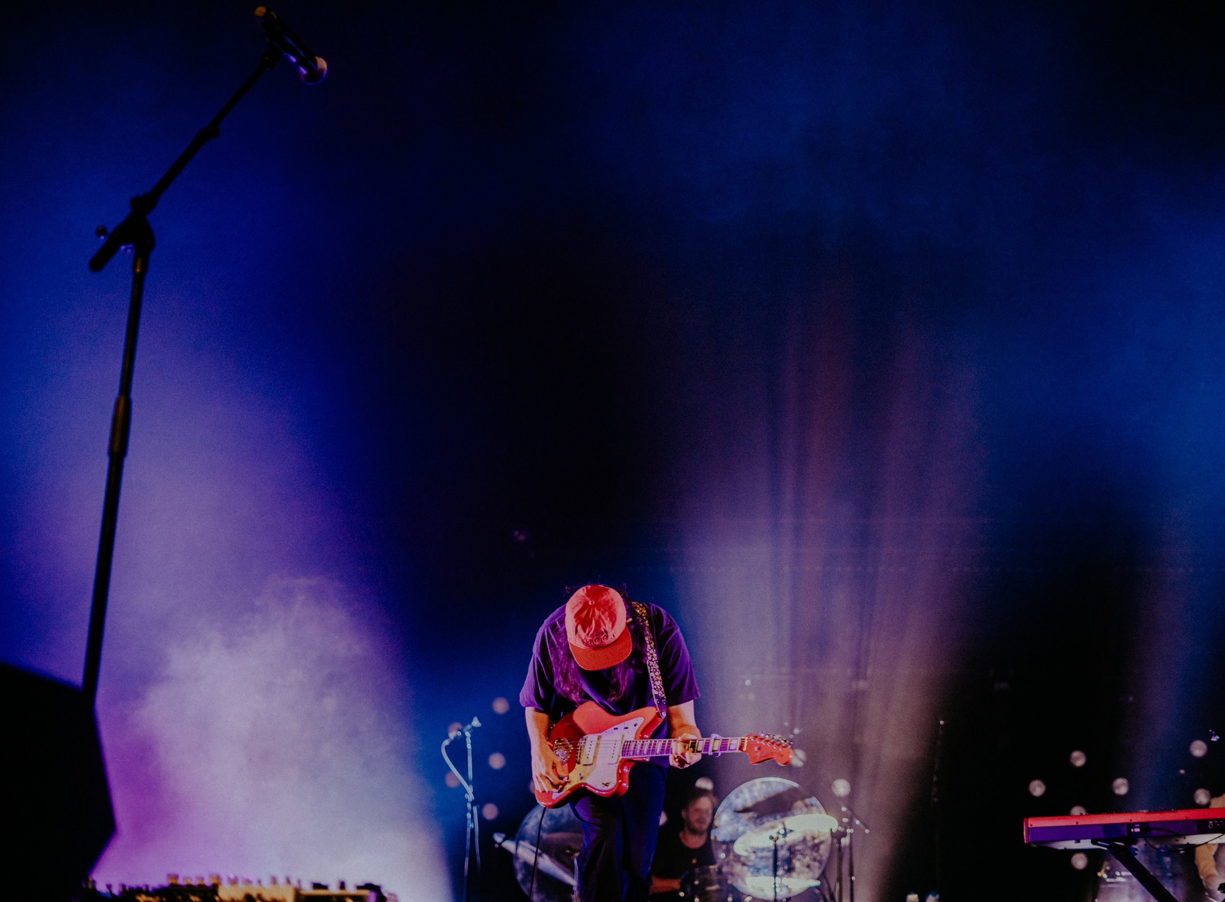 Musician on stage playing an electric guitar with drum set in the background, stage lights illuminating the scene.