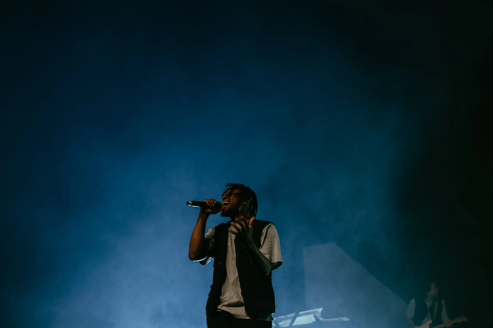 A male singer with dreadlocks performs on stage holding a microphone, with a pianist and guitarist in the background under blue stage lighting.