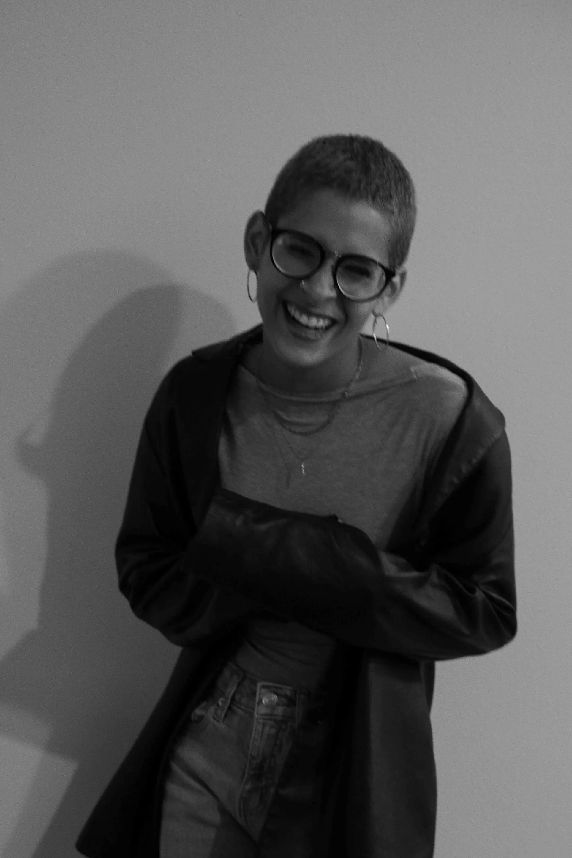 A person with short hair and large glasses, smiling widely, wearing a t-shirt, jacket, and earrings, standing against a plain wall.