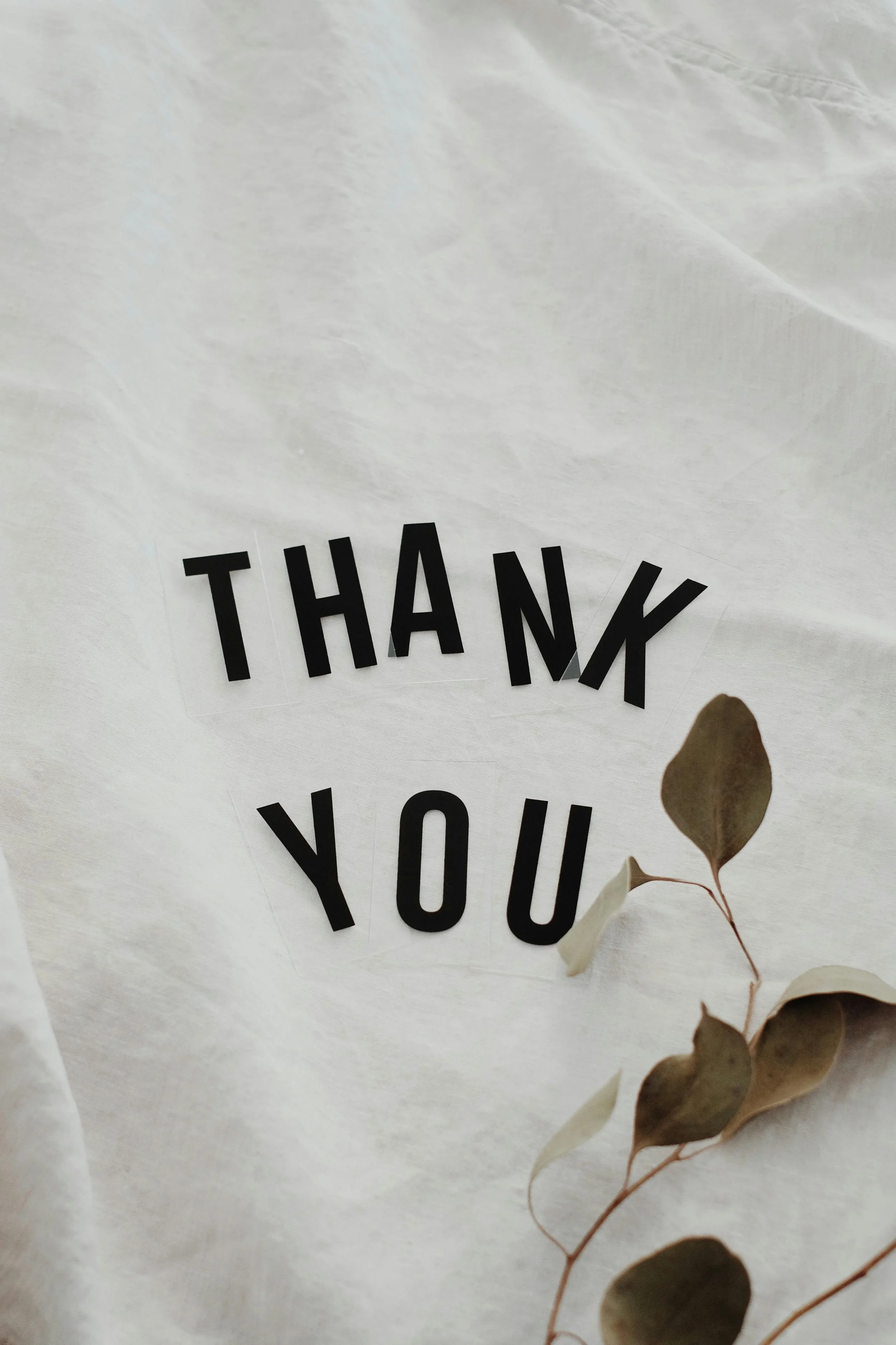 A thank you card with black cut-out letters spelling 'THANK YOU' on a light fabric background, with a sprig of dried eucalyptus leaves in the lower right corner.