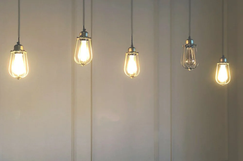 Five hanging Edison-style light bulbs with exposed filaments, lit and suspended from cords in a line against a plain wall.