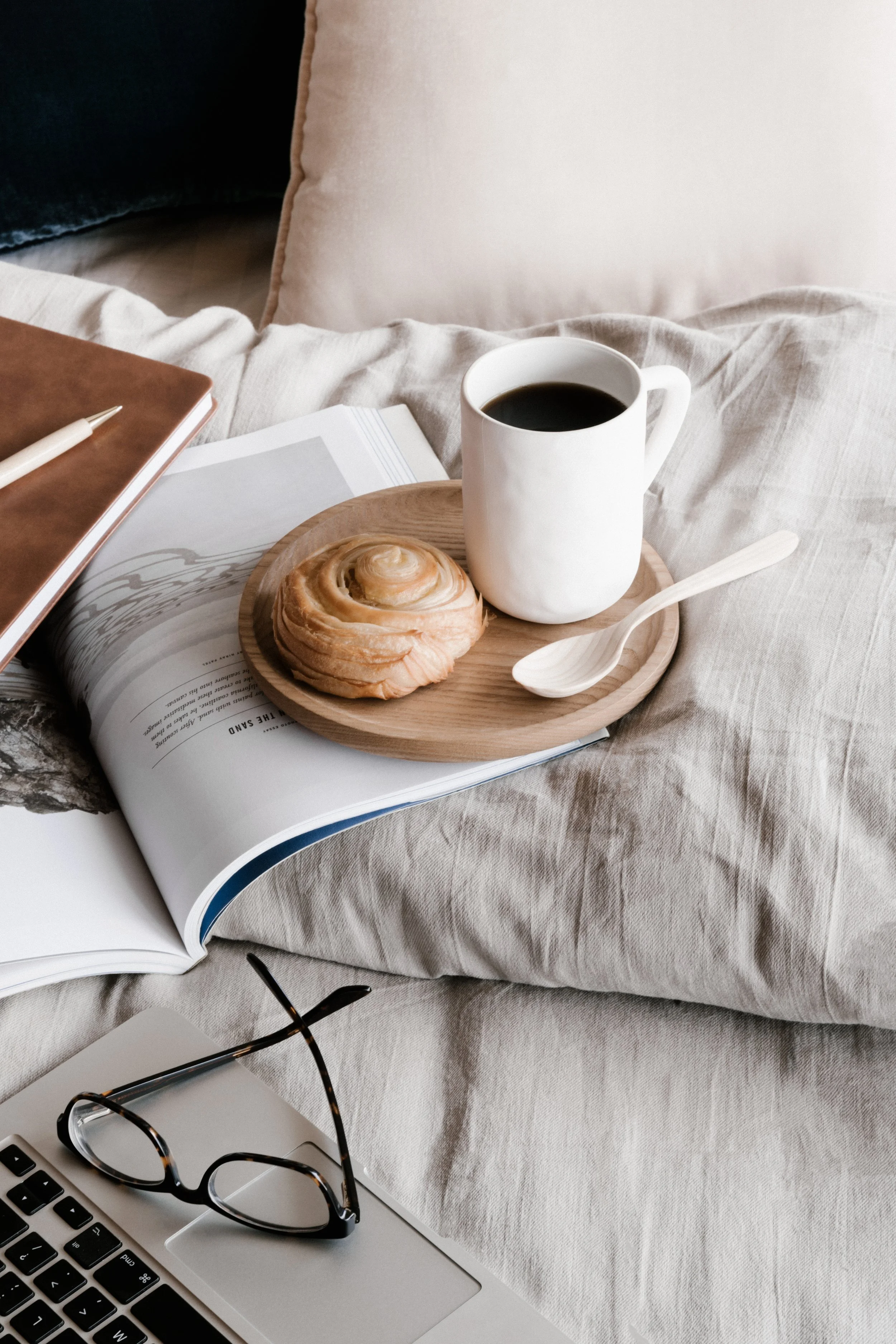 A cozy setup on a bed with a book, a pair of glasses, a notebook, and a tray holding a mug of black coffee, a cinnamon roll, and a wooden spoon.