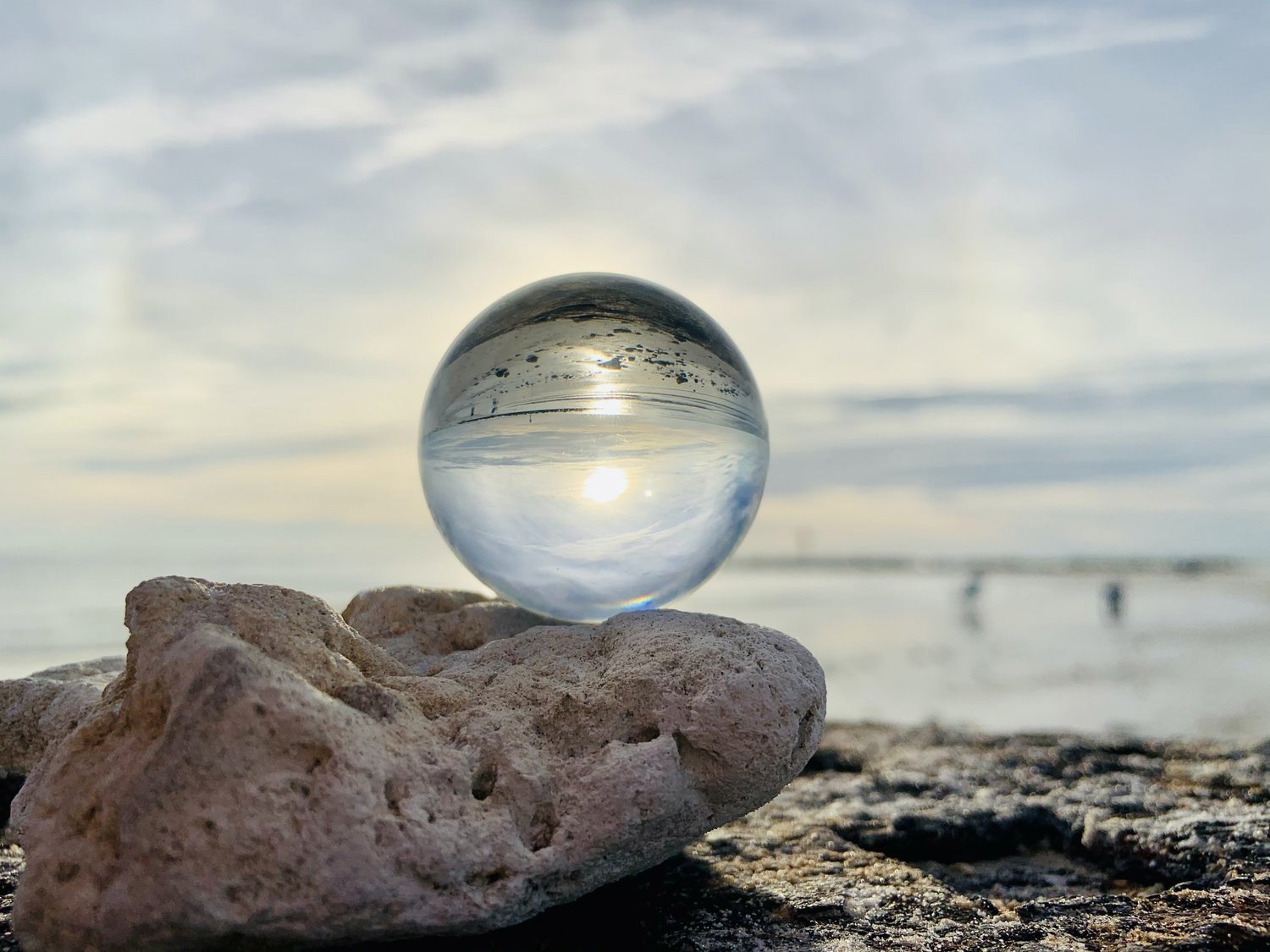 Glass sphere placed on a rocky surface at the beach, reflecting the sky, clouds, and the sun during sunset or sunrise.