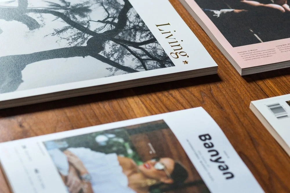 Several magazines laid out on a wooden table, including 'Living,' 'Banyan,' and others, with photo covers of trees, a person, and a woman.