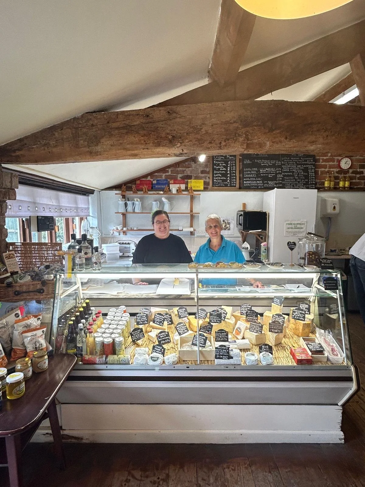 2 x staff are behind the cheese counter smiling. The cheese counter has a glass front and contains lots of cheeses in a neat display. Labels are displayed along side each cheese with a short description, name and price