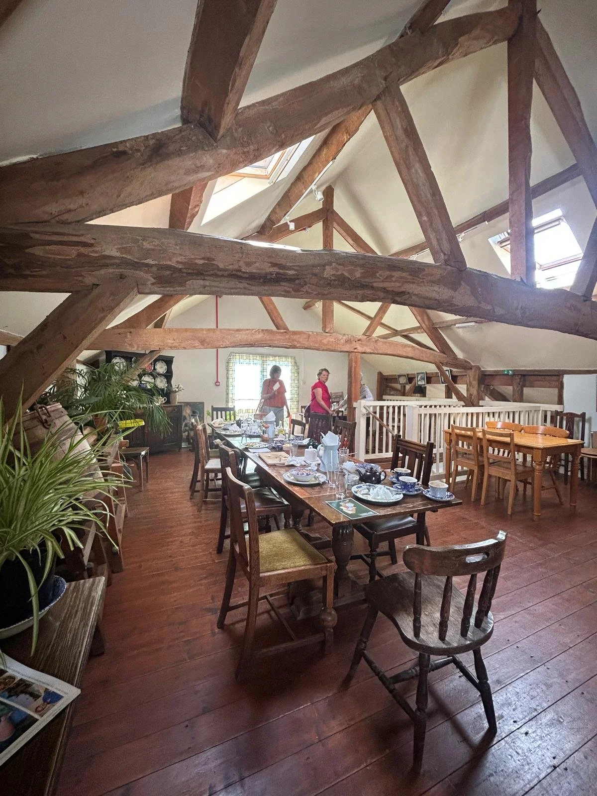 The upstairs of the cafe - showing wooden chairs & tables in a large room with wooden beams on the ceiling. Pot plants are on a table to the left.