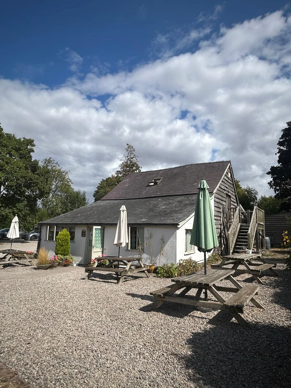 Picture of the outside of the Cafe and Shop - its a white building with a slate roof. Picnic tables with green umbrellas are outside the building on a gravelled area of garden.