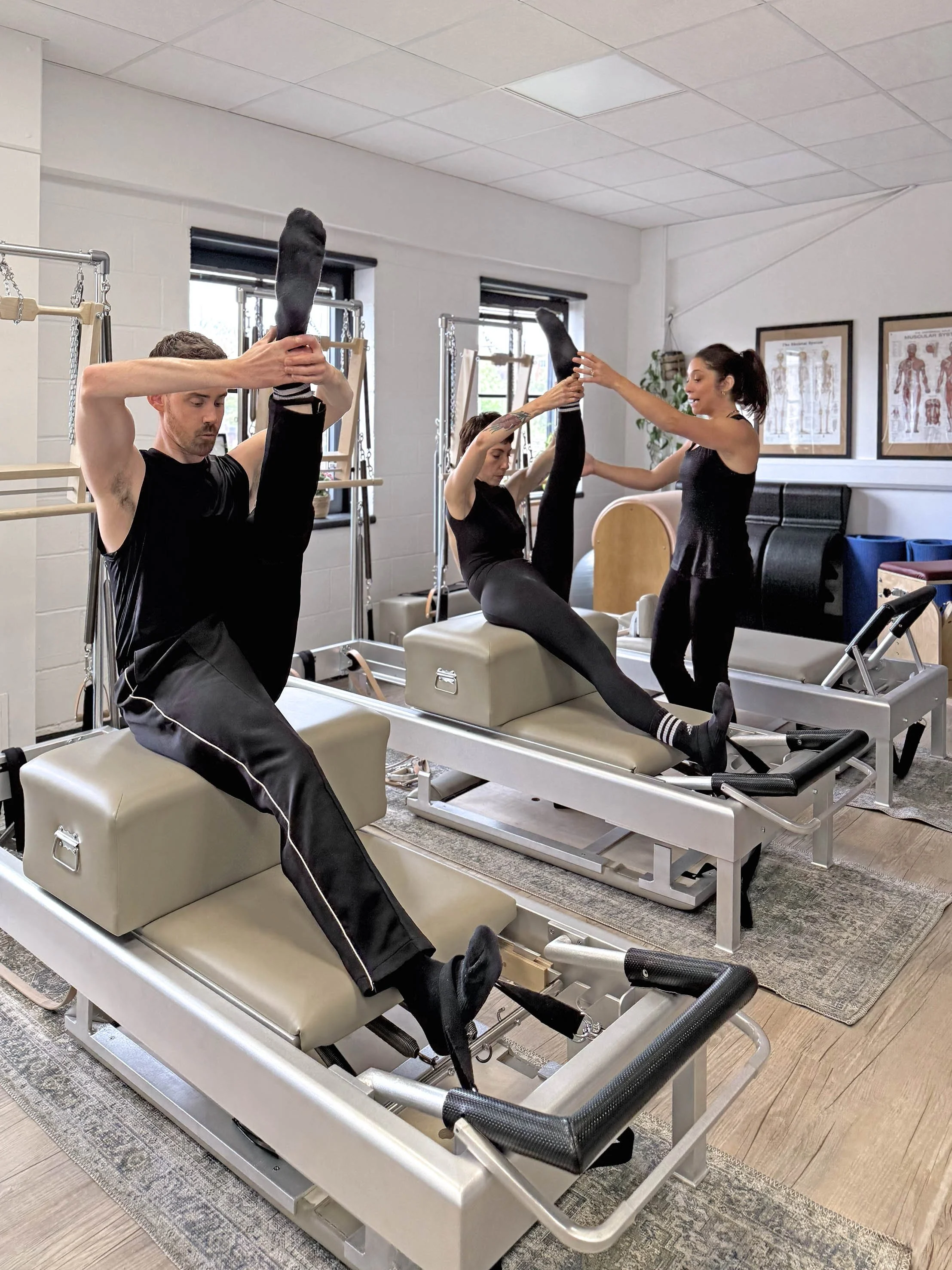 Three people exercising on Pilates reformer machines in a fitness studio with one of our experienced teachers assisting and guiding.