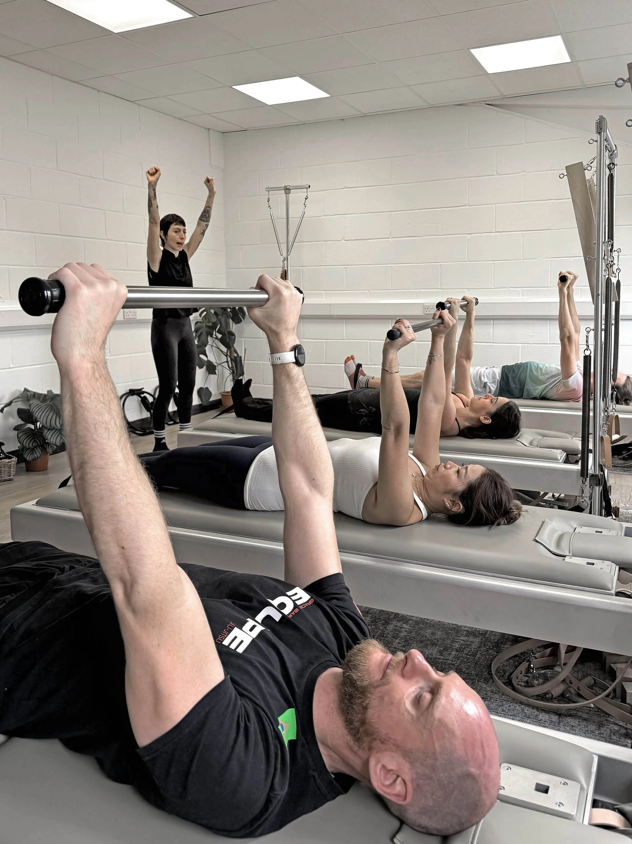 People lying on a classical Pilates mat exercising with bars, with an experienced teacher standing behind them in a studio in Clapham. Guiding them with safety in mind.