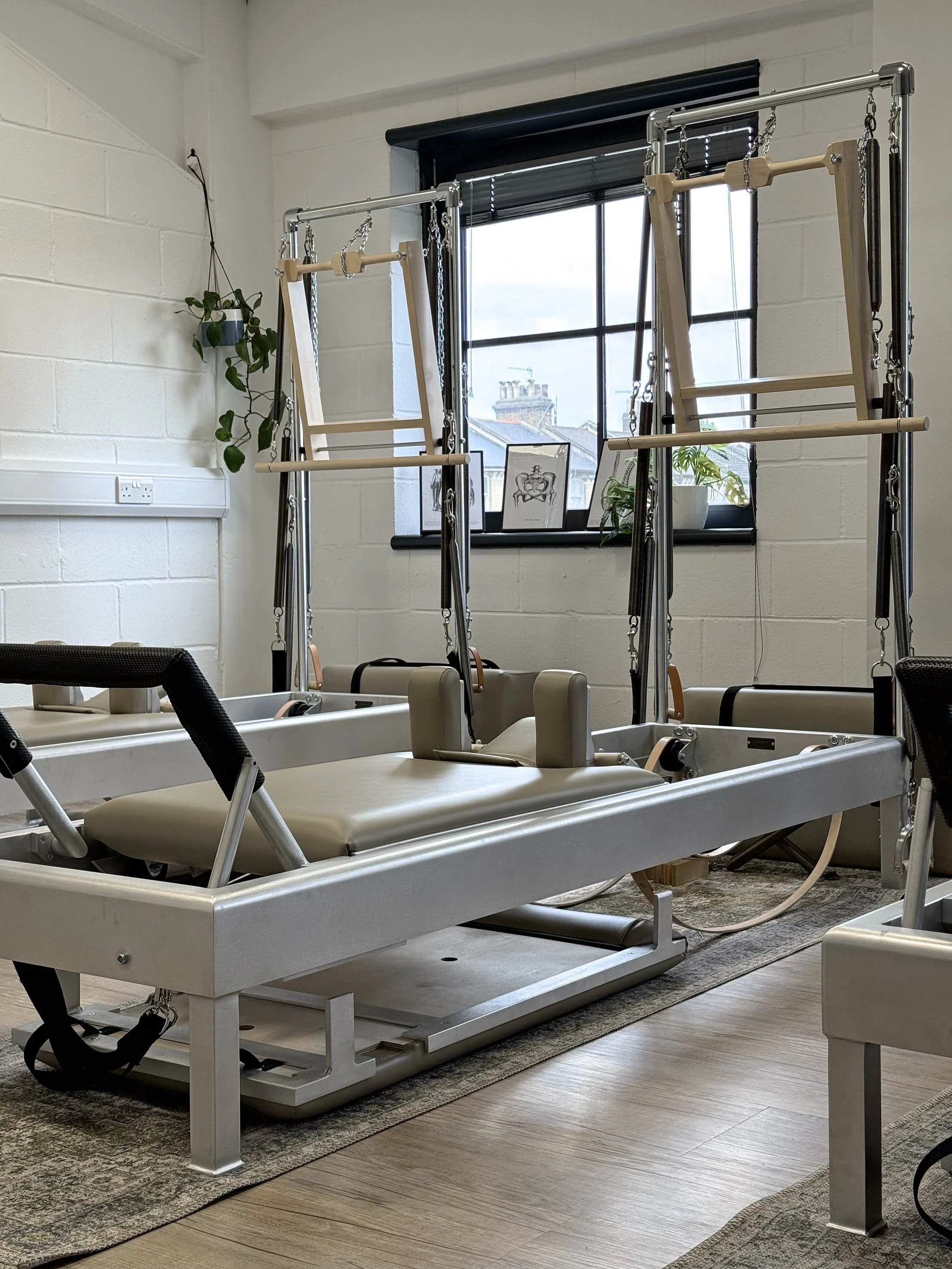 Traditional Classical Pilates equipment including a reformer and two tower tables in a bright room with large window and plants. In the heart of Clapham.