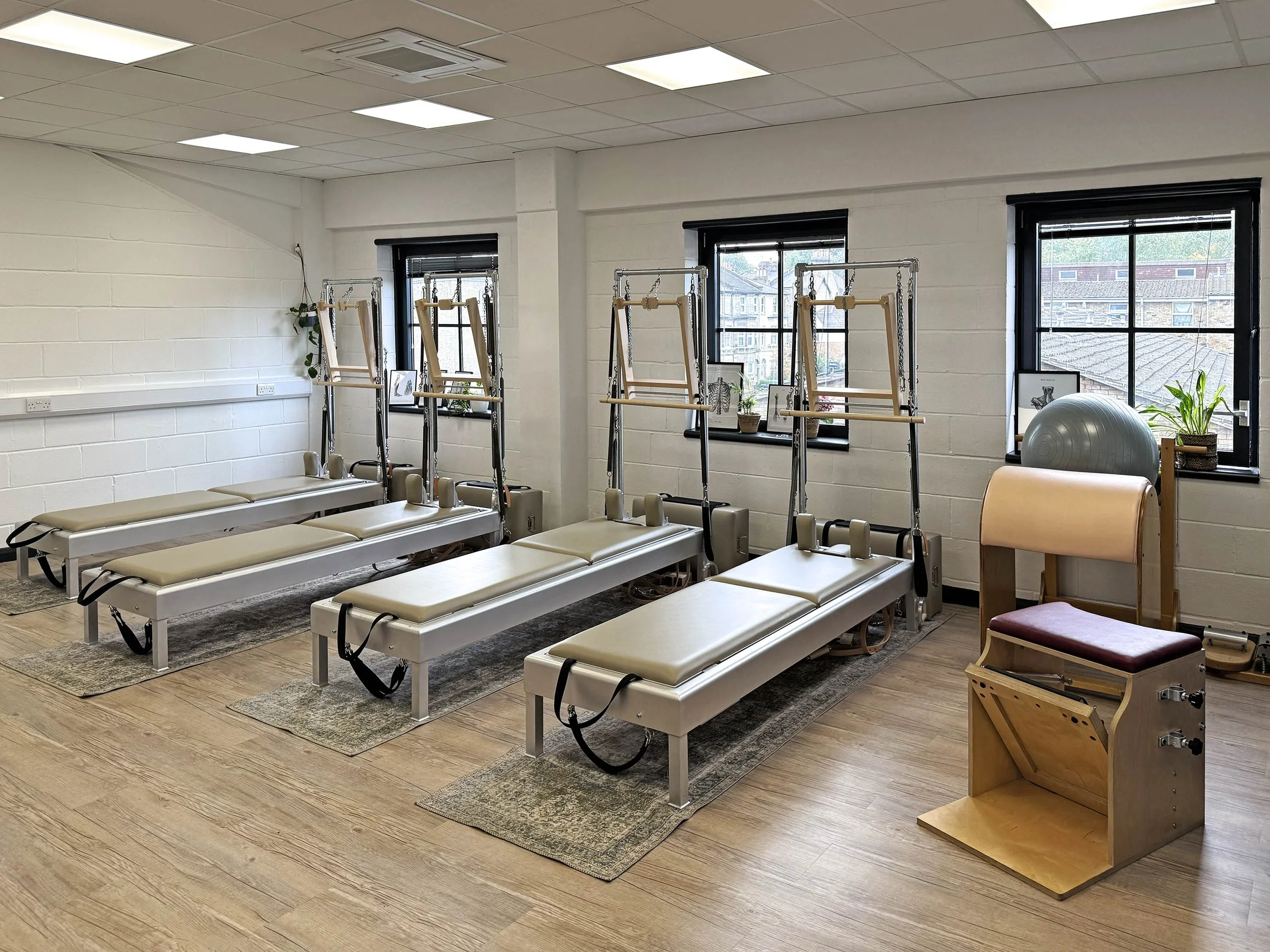 A beautiful airy room with 4 Tower mats, Pilates reformers, a balance ball, and a wooden chair with padded support near windows. The room has white brick walls, wooden flooring, and natural light from three windows. Clapham north